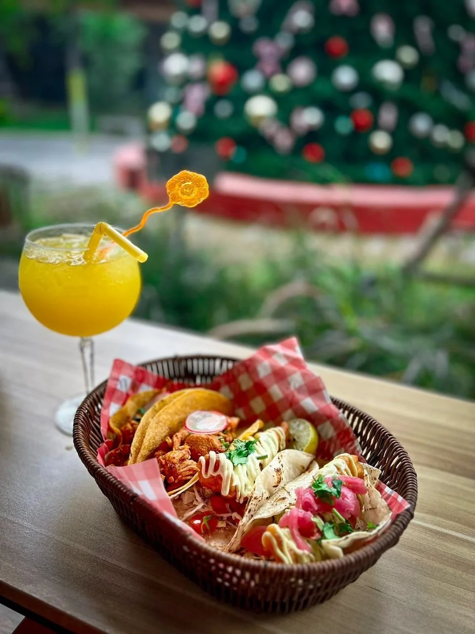 A basket of tacos with various toppings on a wooden table, accompanied by a yellow drink with a flower-shaped garnish and a curly straw, with a blurred background of a decorated Christmas tree outside.