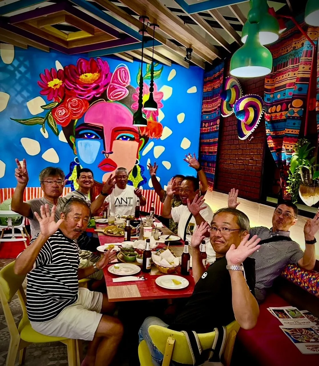 Group of people sitting at a restaurant table waving and smiling, with colorful, artistic wall mural in the background featuring a woman's face with vibrant flowers and bold colors.