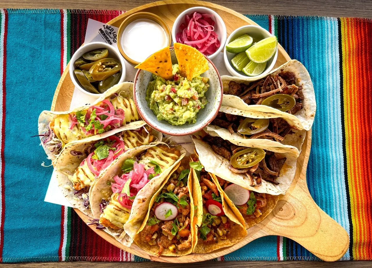 A wooden serving tray with six tacos, each filled with various meats and toppings, accompanied by lime wedges, pickled onions, sliced jalapenos, guacamole, and small bowls of sour cream, sauce, and additional toppings, on a colorful woven tablecloth.