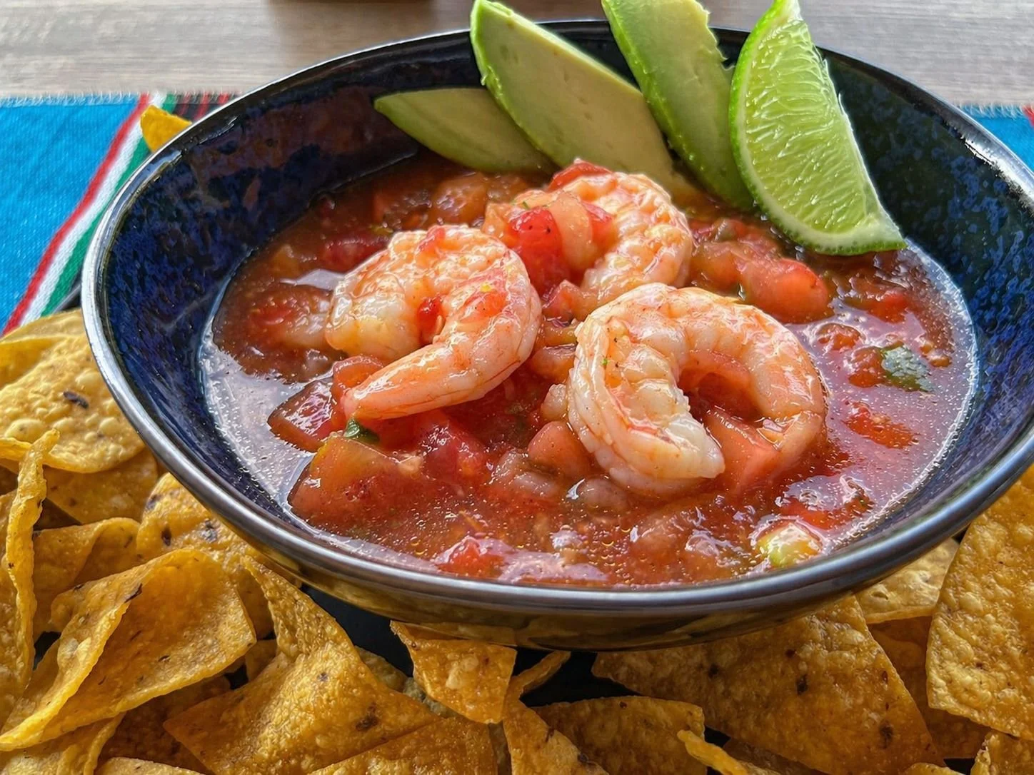 A bowl of shrimp ceviche garnished with lime wedges. Corn tortilla chips are placed around the bowl on a colorful striped cloth.