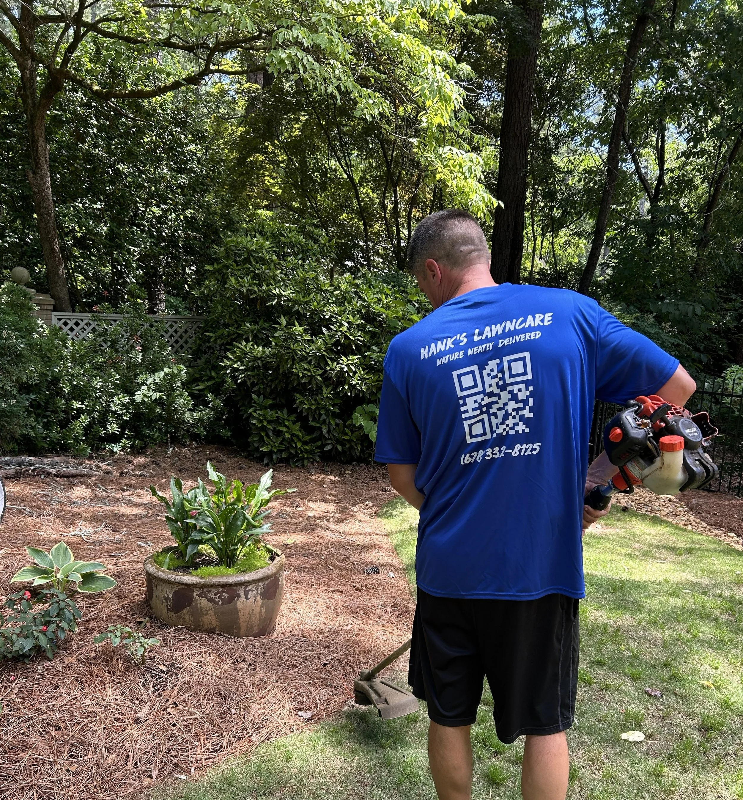 A man wearing a blue shirt with white text on the back, including a QR code, is working outside in a garden or backyard surrounded by dense green trees and plants. The man is holding a gardening or landscaping tool and is standing on a patch of grass with mulch and potted plants nearby.