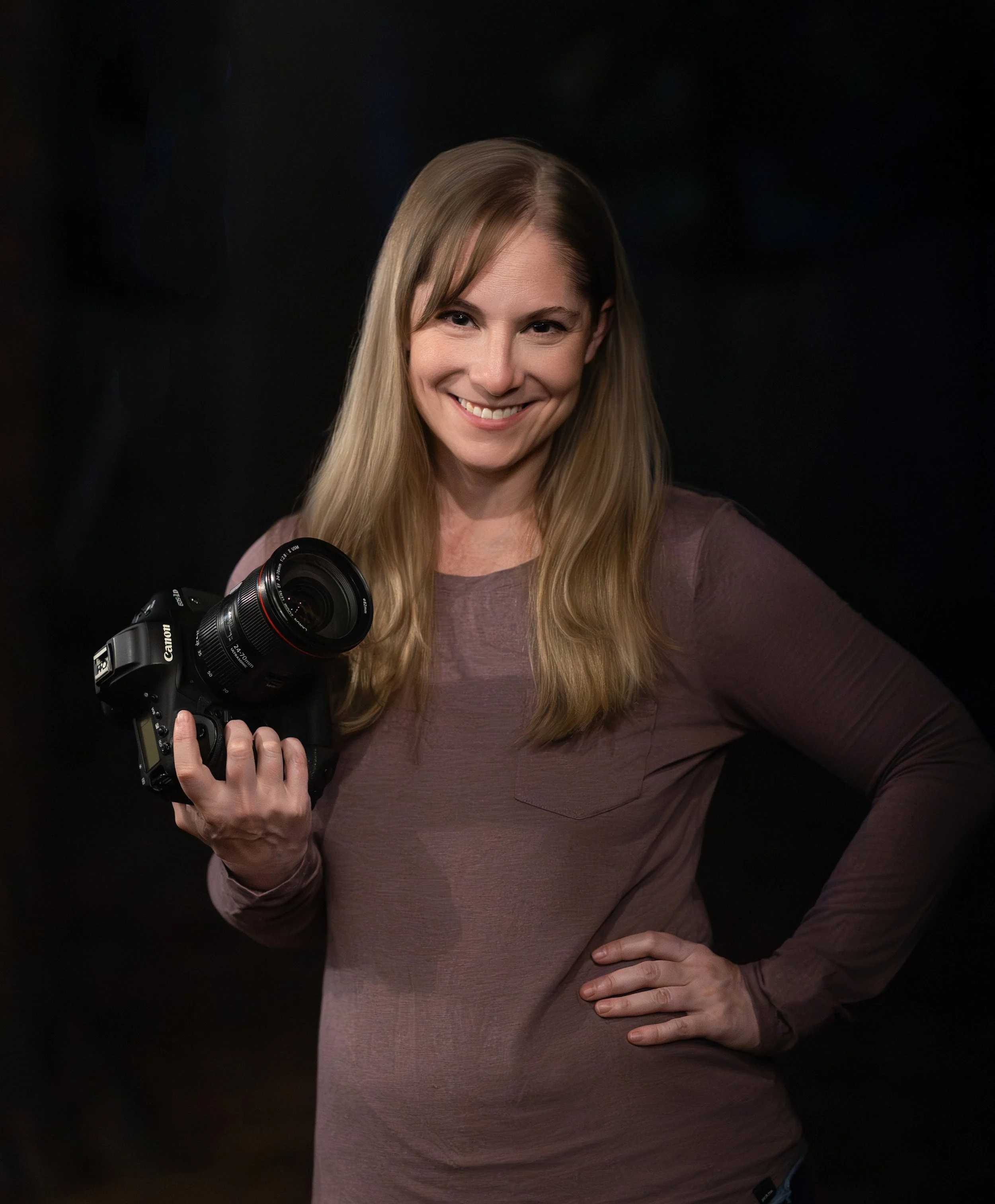 A light-skinned woman with long hair smiles while holding a camera. She wears a long-sleeved shirt and appears confident.