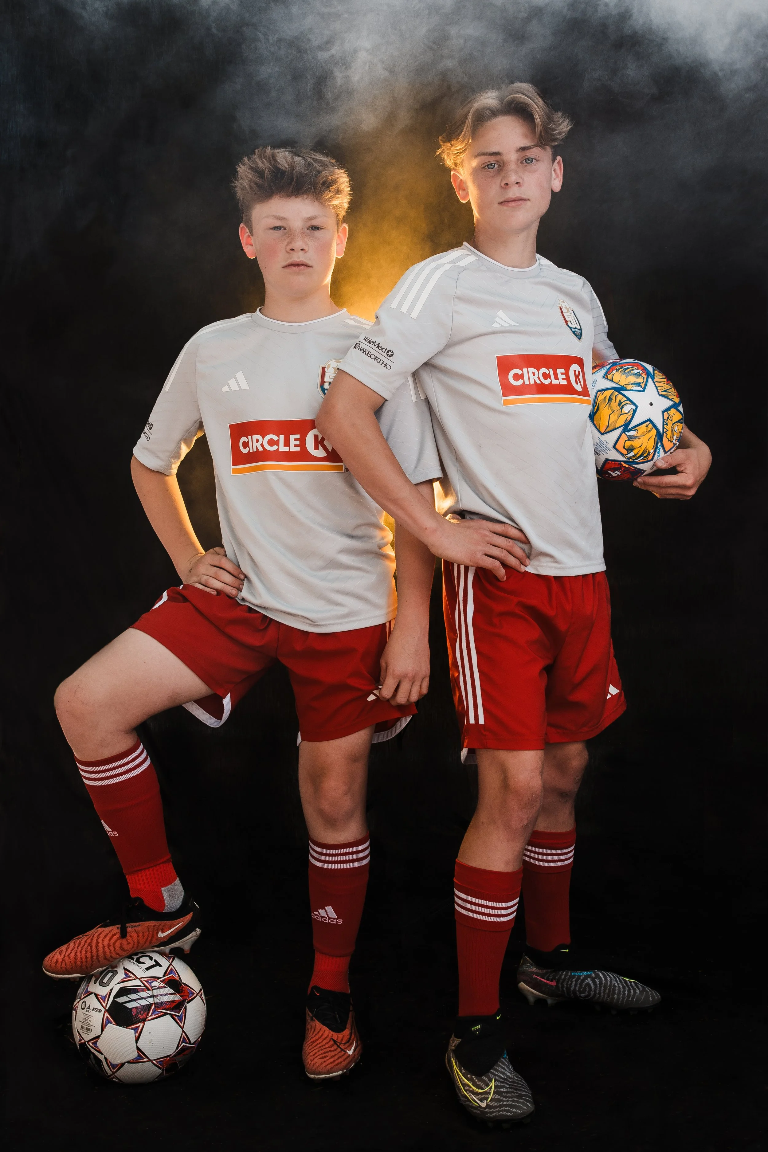 Two teenage boys in soccer uniforms pose confidently against a dark background. One has his foot on a soccer ball, and the other holds a ball by his side. Both wear white shirts, red shorts, and red socks, with a yellow light behind them.