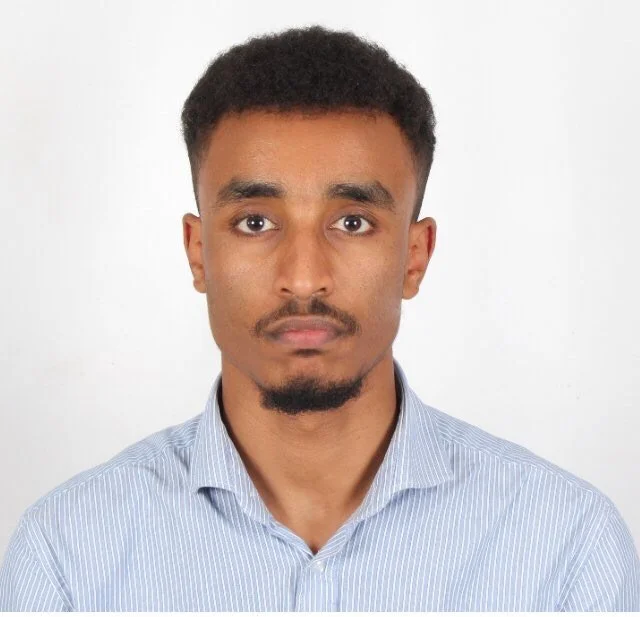 Portrait of a young man with short curly hair and a trimmed goatee, wearing a light blue striped shirt, against a plain white background.