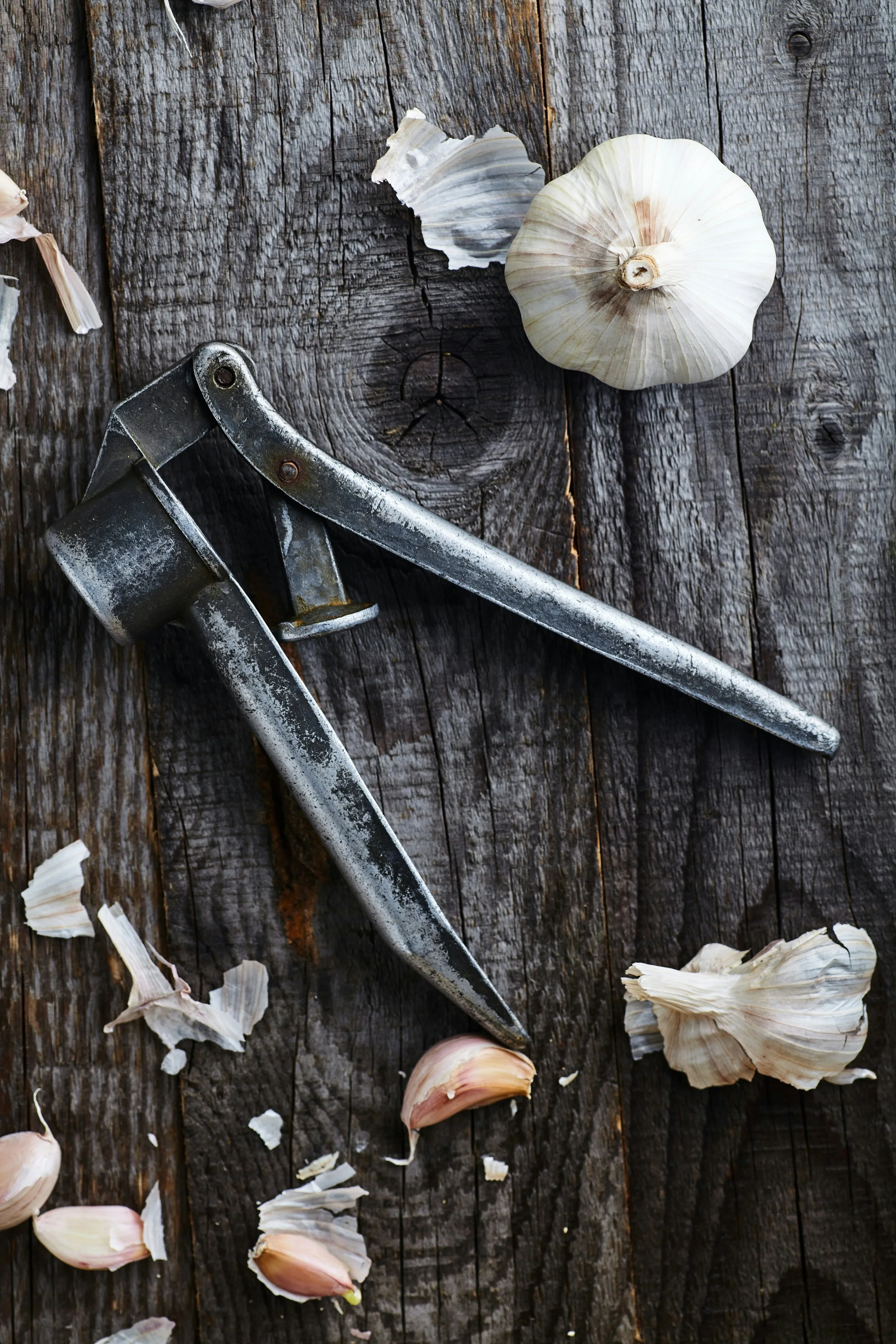 A weathered wooden surface with garlic bulbs, garlic skins, and peeling garlic cloves scattered around, along with a metal garlic peeler or tool.