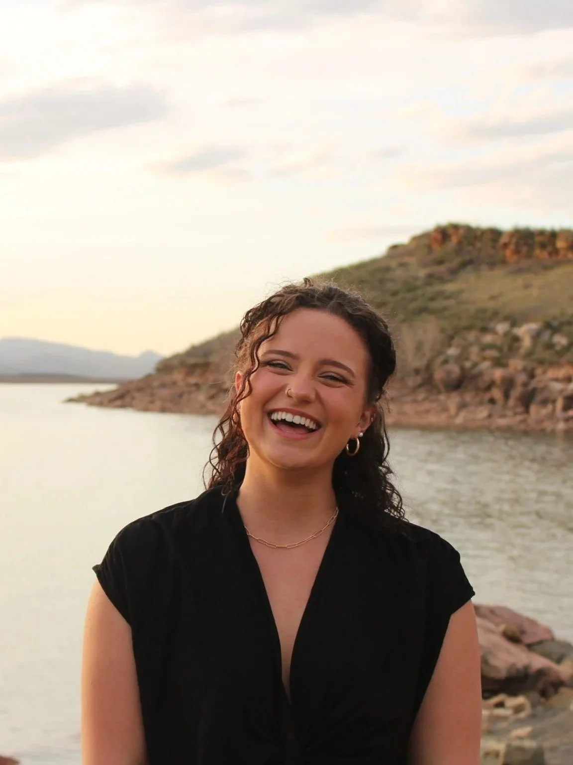 A woman with curly hair smiling outdoors near a body of water and rocky hills in the background during sunset.
