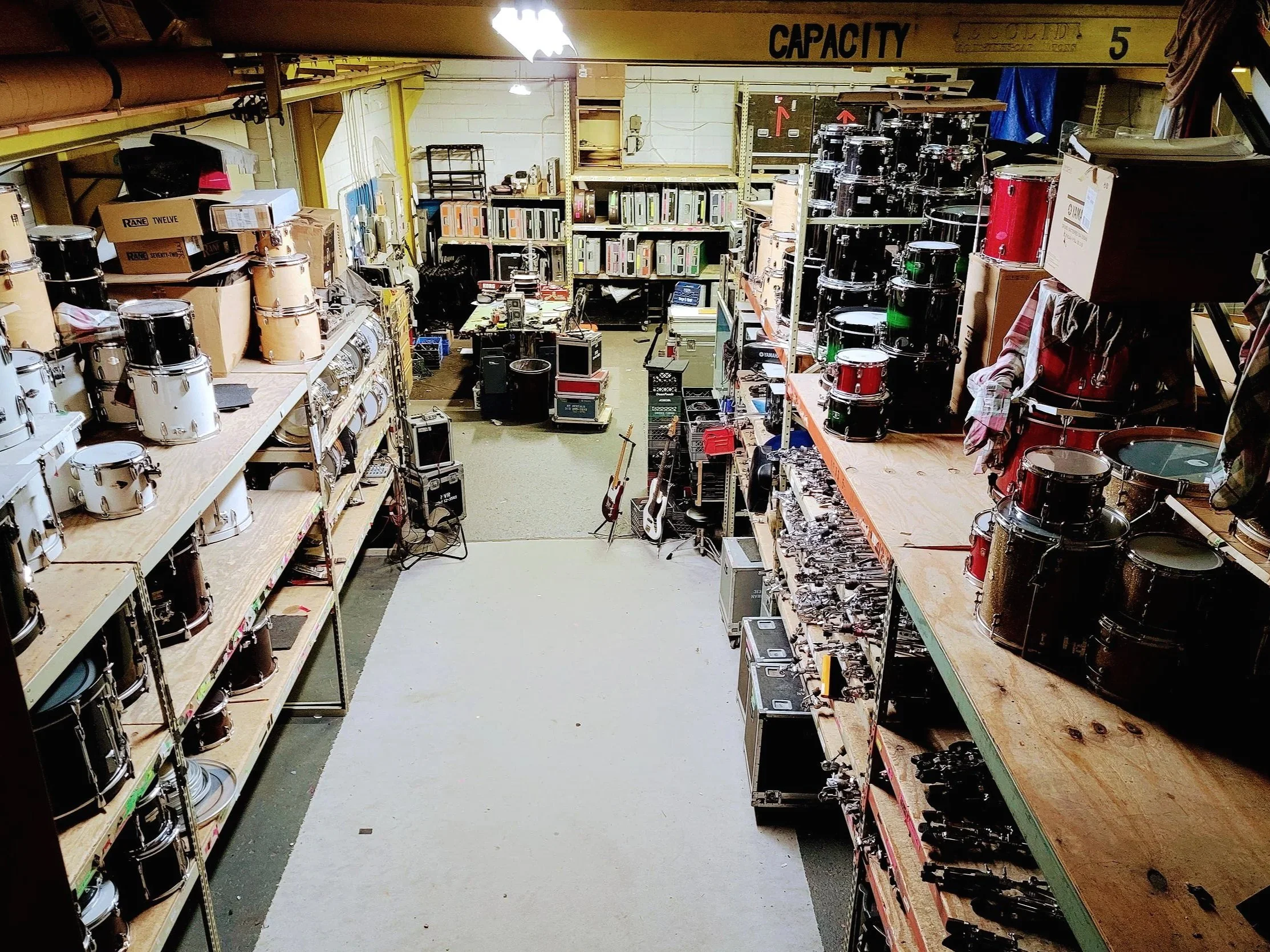 A storage room filled with drum equipment and accessories, including drums on shelves, guitar cases, and other musical gear.