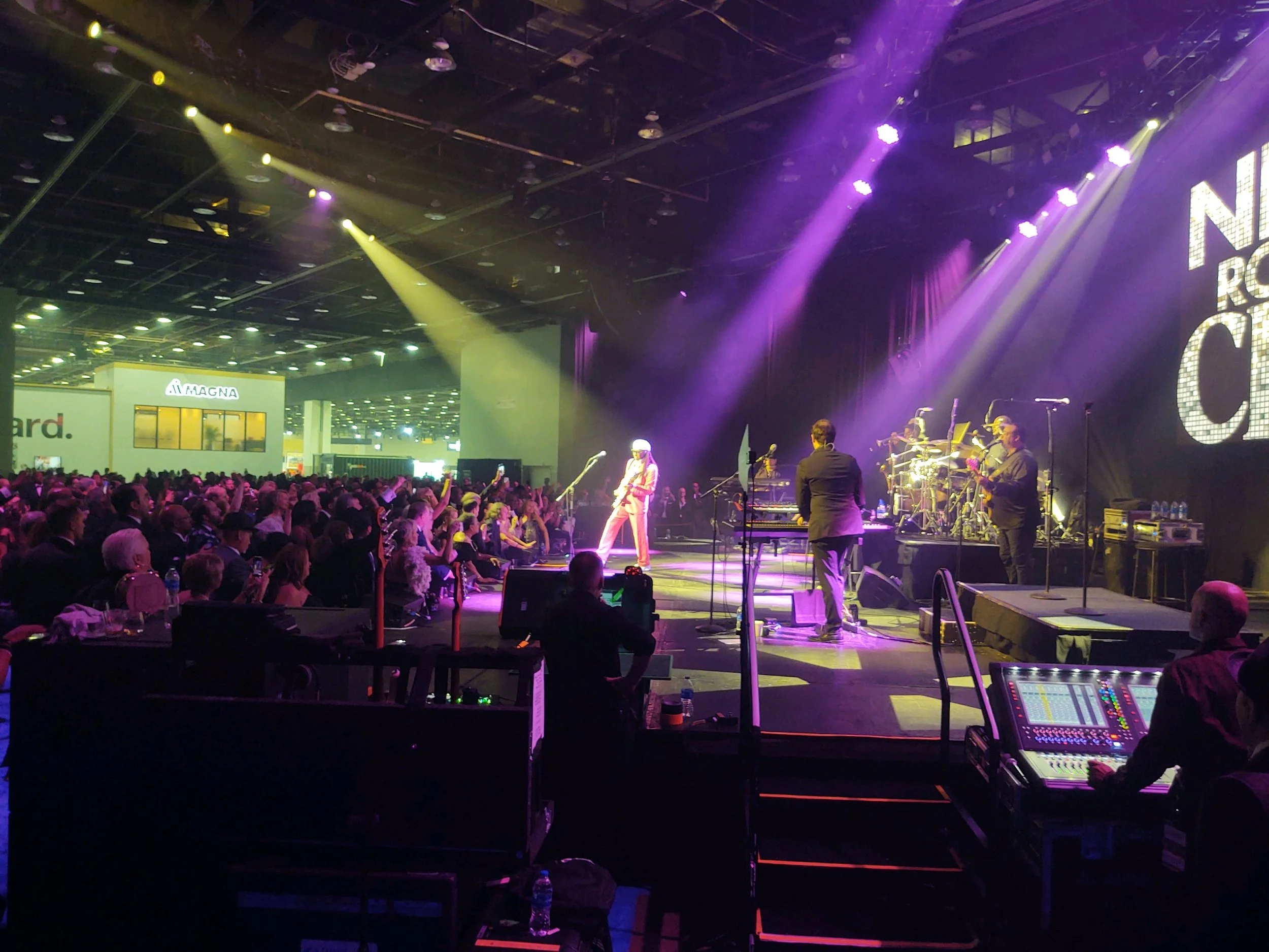 Stage with band performing at a concert, audience watching, colorful lights illuminating the scene.