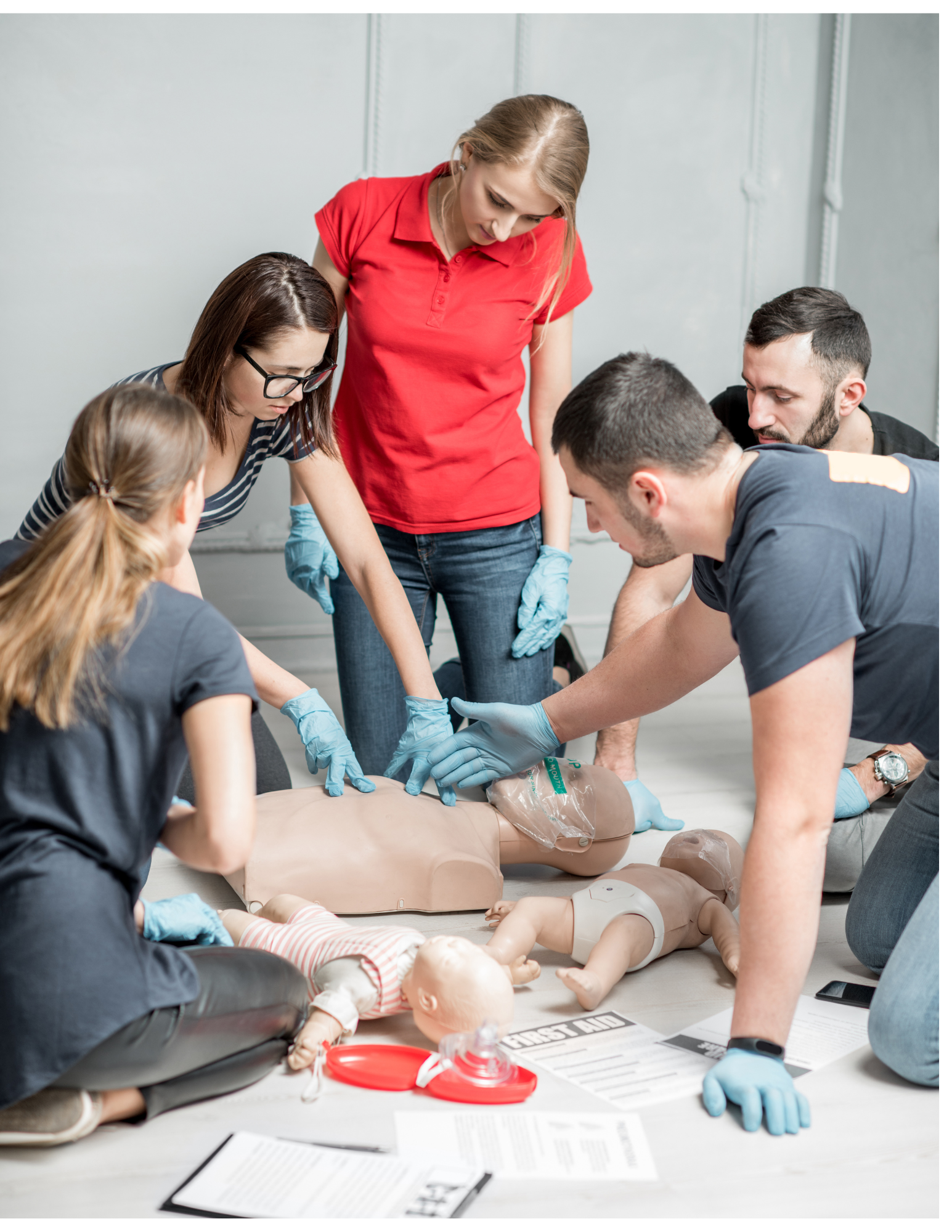 A CPR training session with a group of people practicing on mannequins, led by an instructor in a red shirt. The trainees are wearing gloves and are focused on the hands-on procedure.