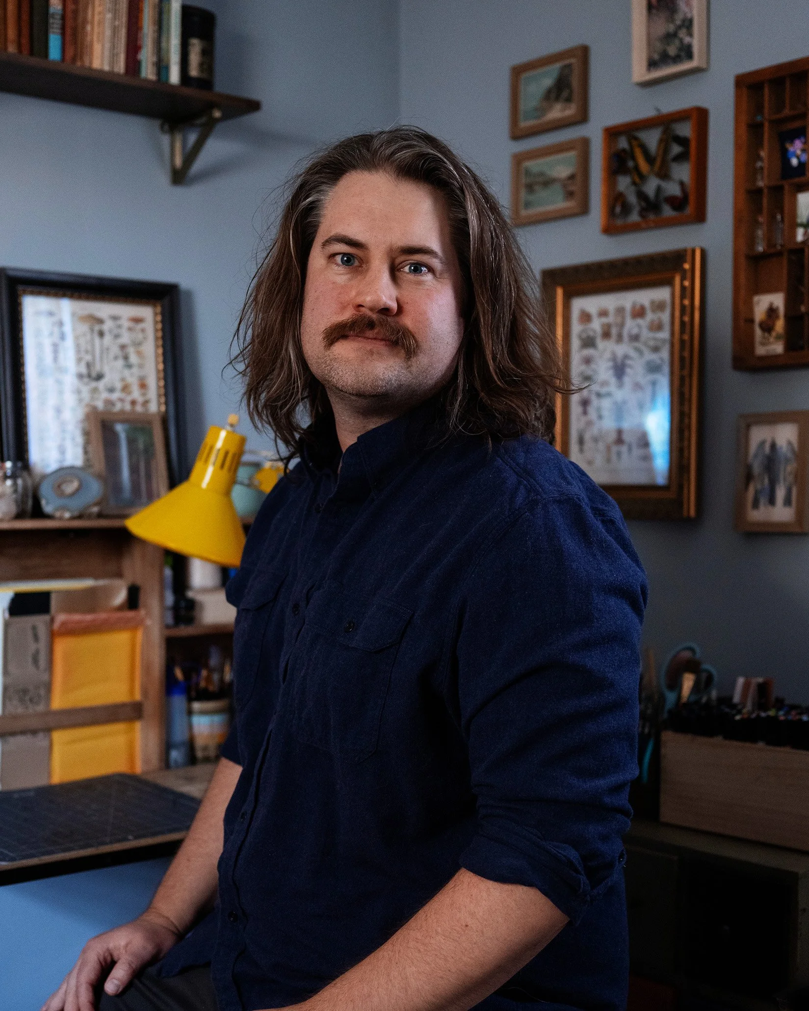 A portrait of Ryan Lynch in his work space. He has long hair and a mustache sitting in a room with framed pictures and books on shelves, wearing a dark blue shirt.