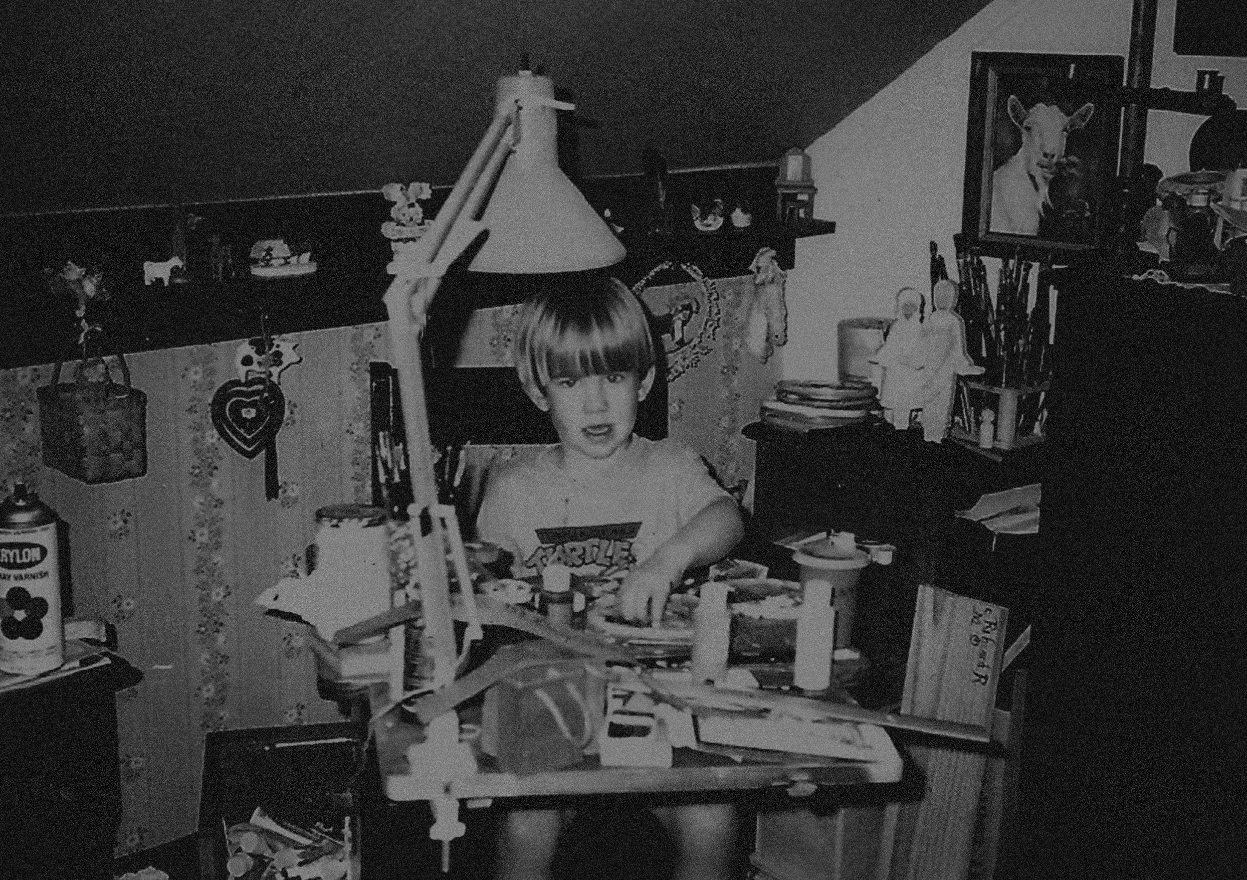 A 1990s photo of Ryan Lynch sitting at a cluttered painting desk in a room with various objects, including a lamp, paintings, and shelves filled with items.