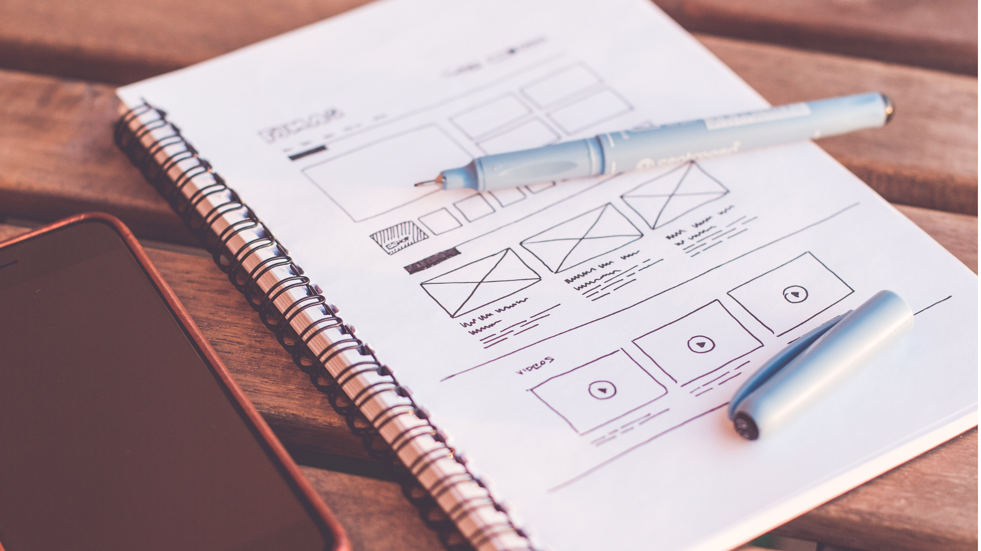 Design sketch of a website layout on white paper with sketches of video thumbnails, text boxes, and interface elements, placed on a wooden desk with a pen and a smartphone.
