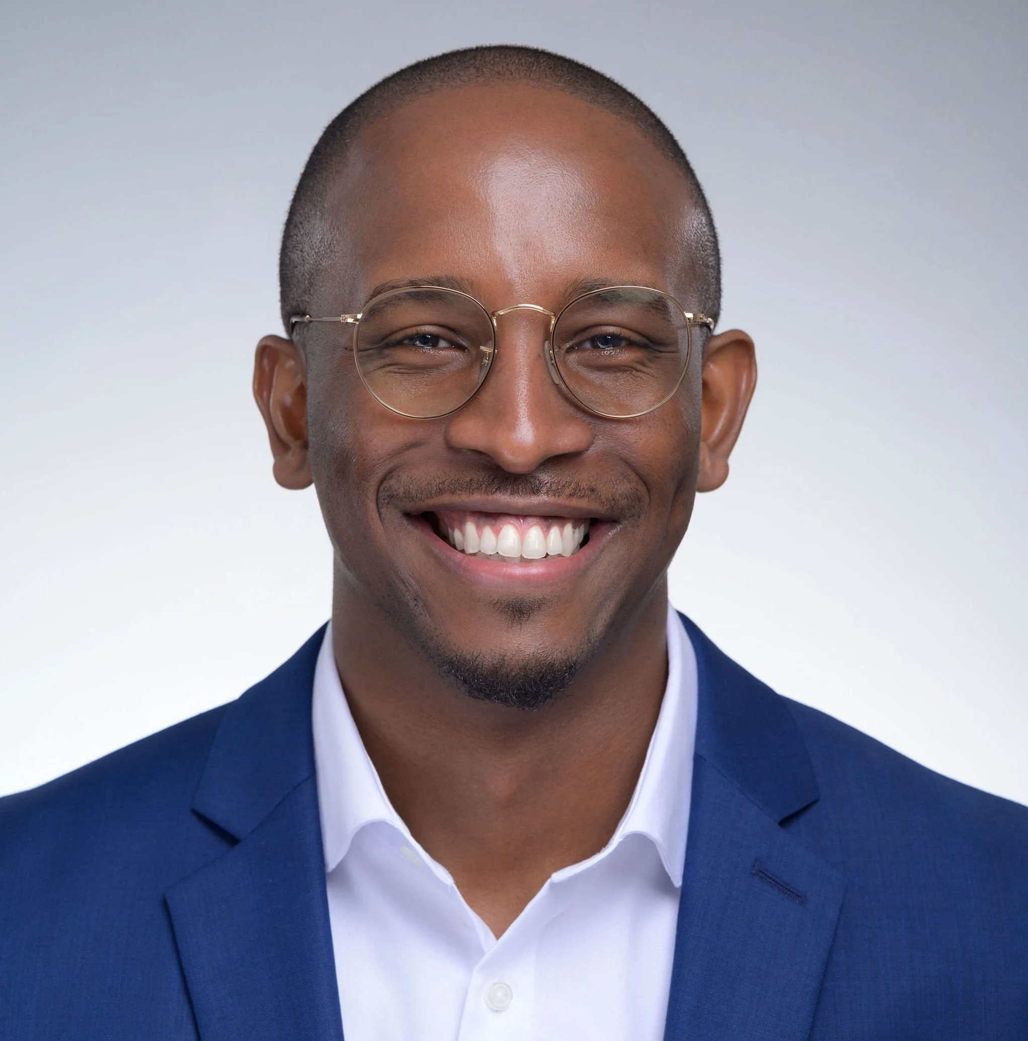 Close-up of a smiling man wearing glasses, dressed in a white shirt and a blue blazer, with a plain light gray background.