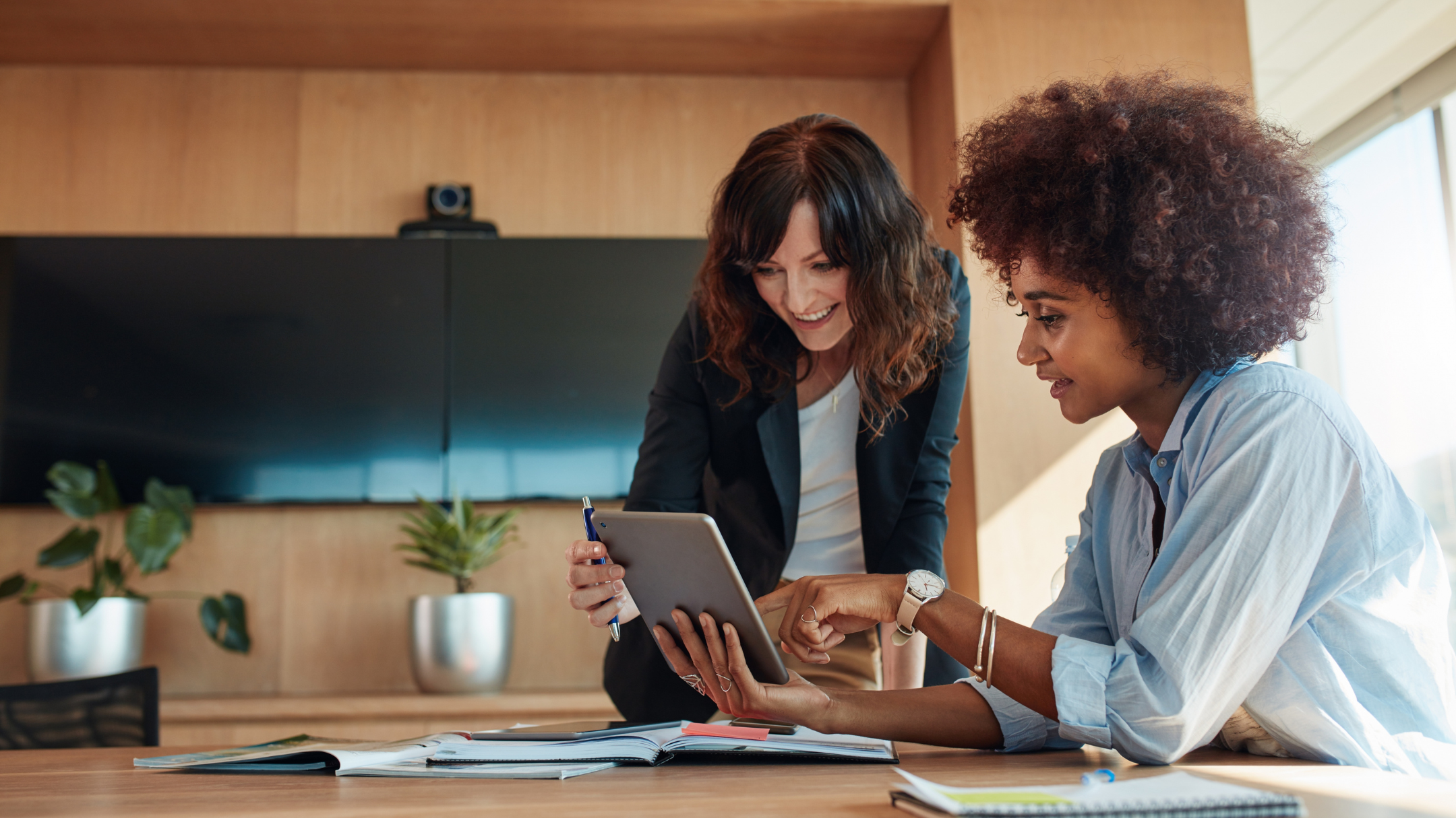 Two women in a modern office, one showing something on a tablet to the other, surrounded by open notebooks and plants.