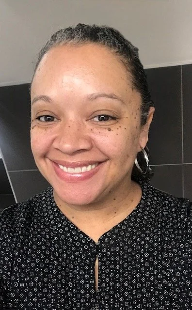 Close-up of a woman with curly hair tied back, smiling, wearing a black patterned top, standing in front of a dark tiled wall.