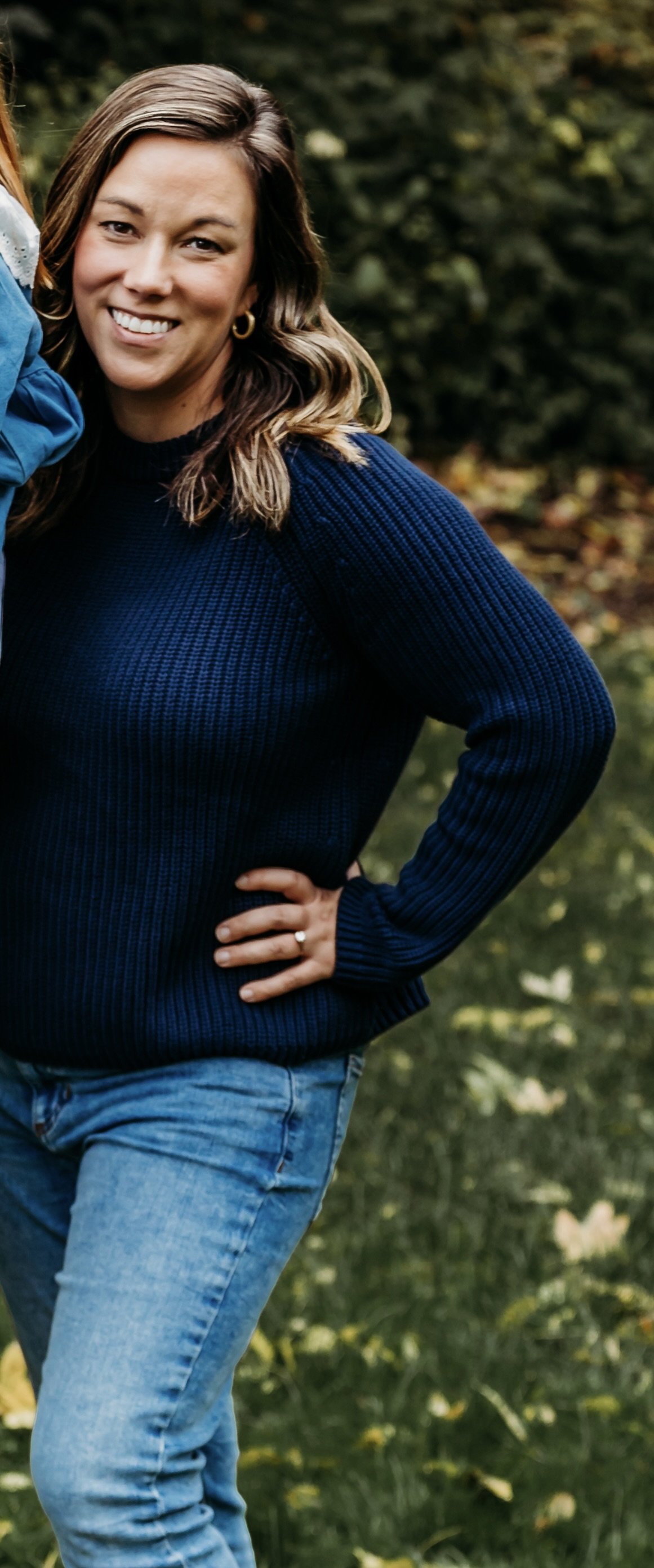 A woman with shoulder-length light brown hair, wearing a navy blue sweater and blue jeans, smiling outdoors in a natural, green setting.