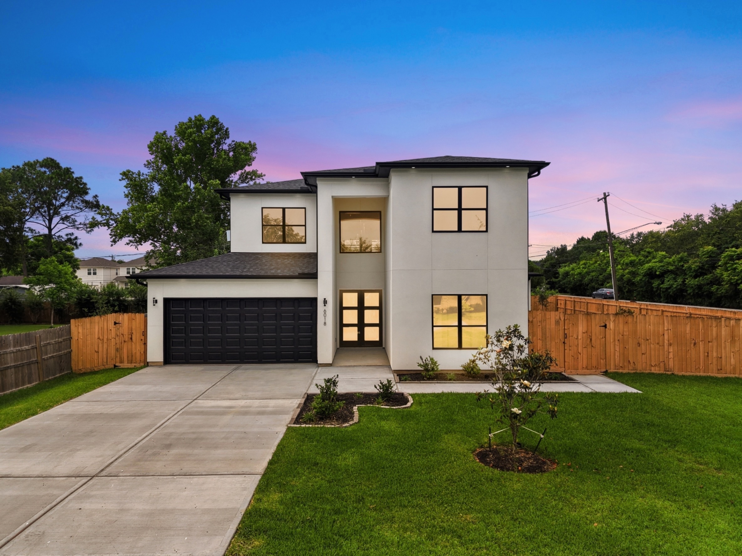 Modern two-story house with a black garage door, large windows, a small front porch, a concrete driveway, green grass, small trees, wooden fence, and power lines against a sunset sky.