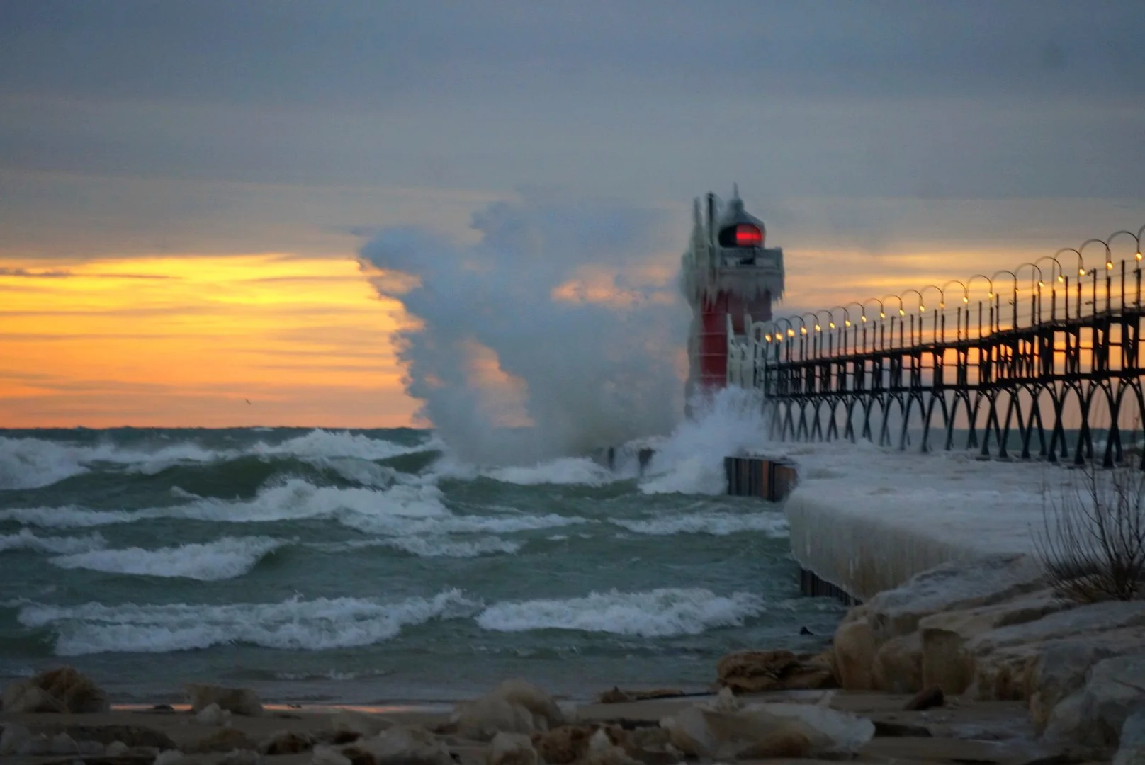 The South Haven pier light glows red at sunset as winter spray bursts beside it.