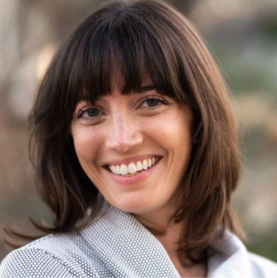 Close-up of a smiling woman with shoulder-length brown hair and bangs, wearing a light-colored blazer.
