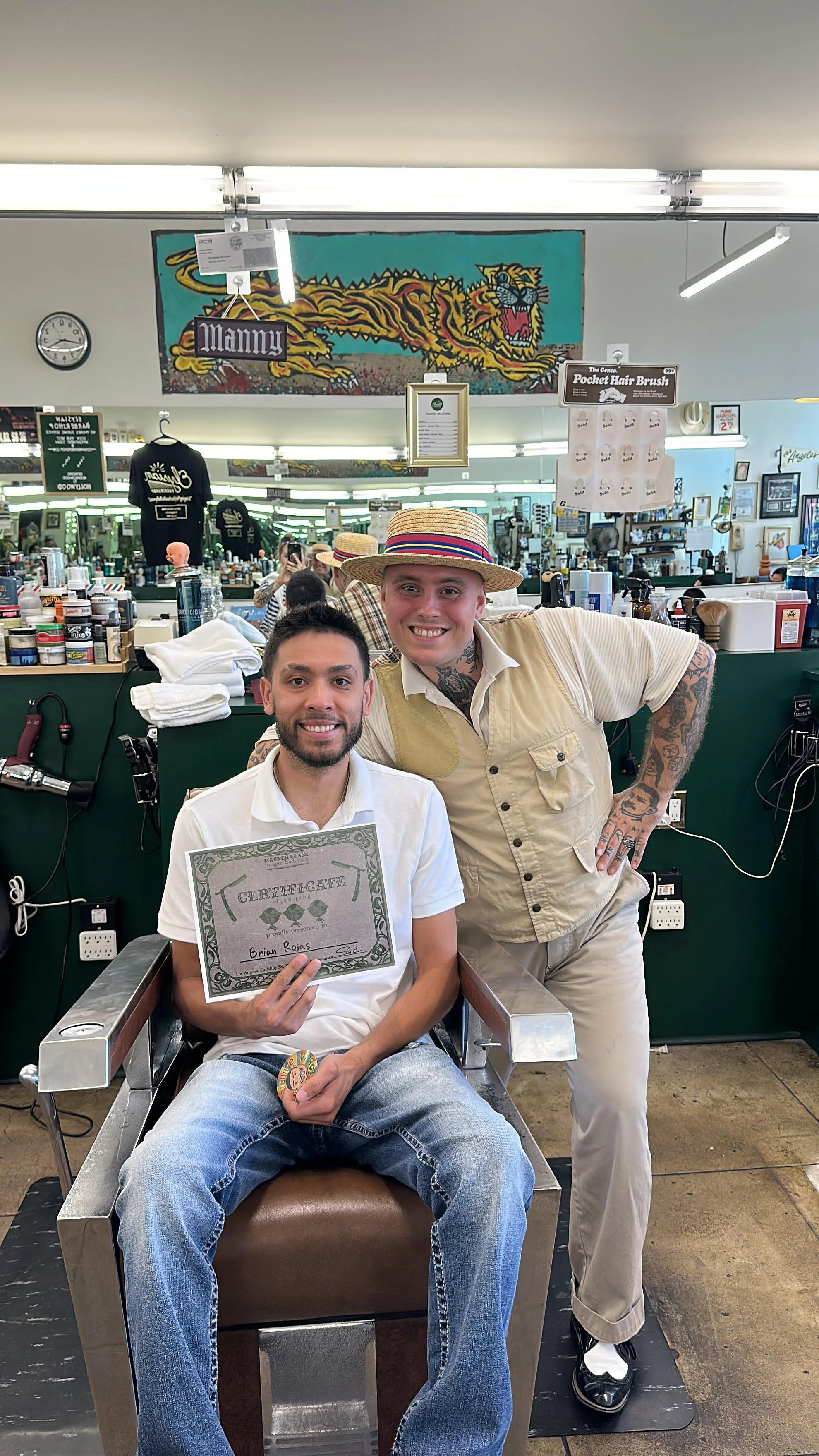 Two men in a barbershop. One man with a certificate around his neck is seated in a barber's chair, holding a certificate and a coin, smiling. The other man stands beside him, leaning slightly, with tattoos on his arms, smiling, wearing a hat and casual clothing. The background shows mirrors, shelves with barber supplies, and various decorations.