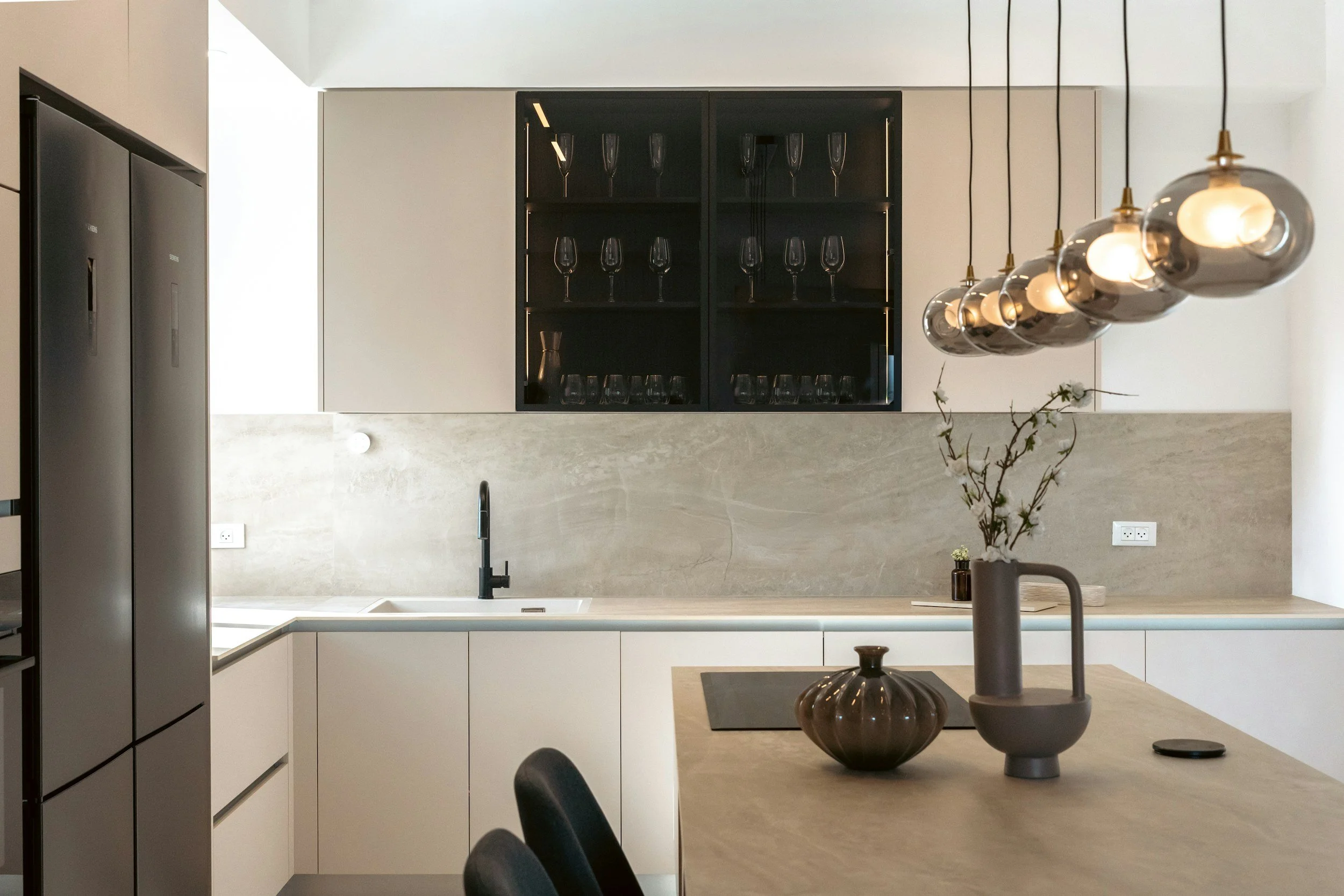 Modern kitchen with beige countertops, white cabinetry, black shelving with glassware, and modern pendant lights with glass globes.