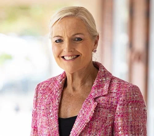 A smiling woman with short blonde hair wearing a pink blazer standing indoors.