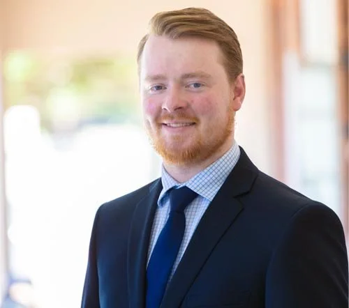 Professional portrait of a young man with red hair and beard, wearing a dark suit, light blue shirt, and blue tie, standing indoors with blurred windows in the background.