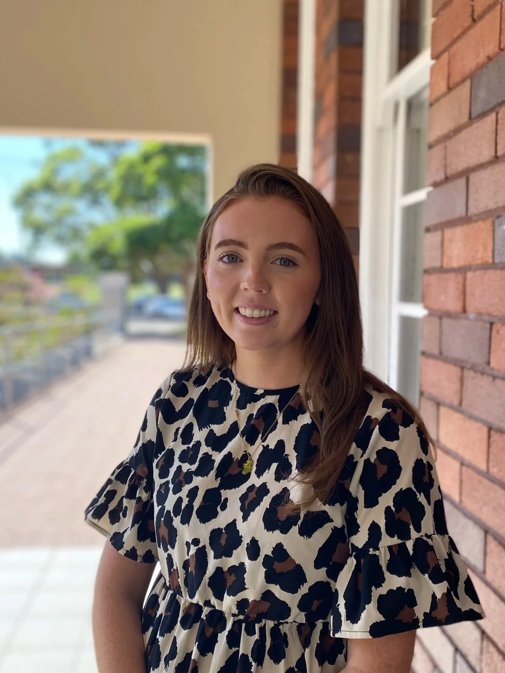 A woman with long brown hair, wearing a black and white leopard print dress, smiling, standing outside a building with a brick wall and large windows, blurred outdoor scenery in the background.