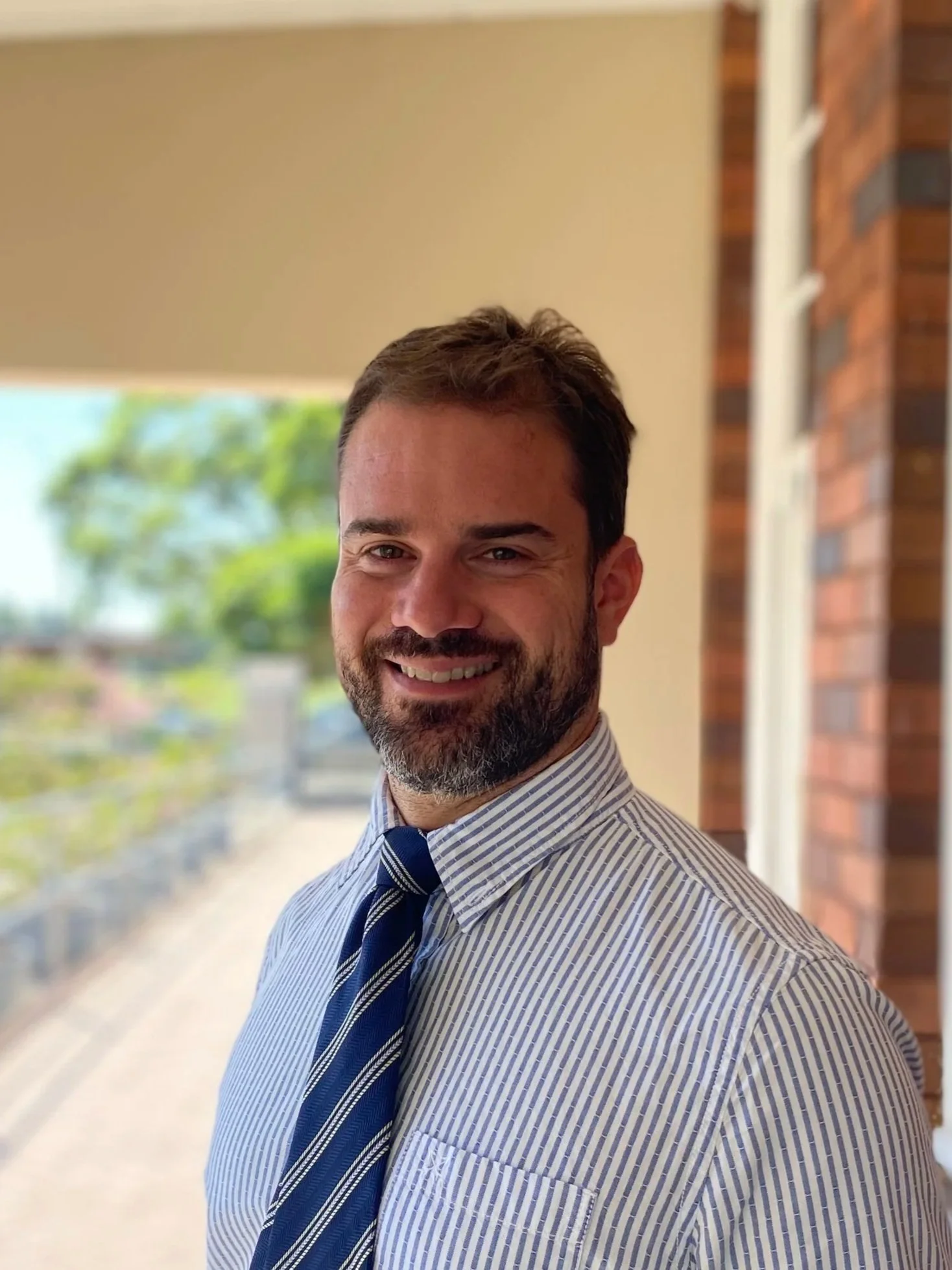 A smiling man with a beard and brown hair, wearing a striped dress shirt and a navy blue striped tie, standing in front of a window with trees outside, and a brick wall to his right.