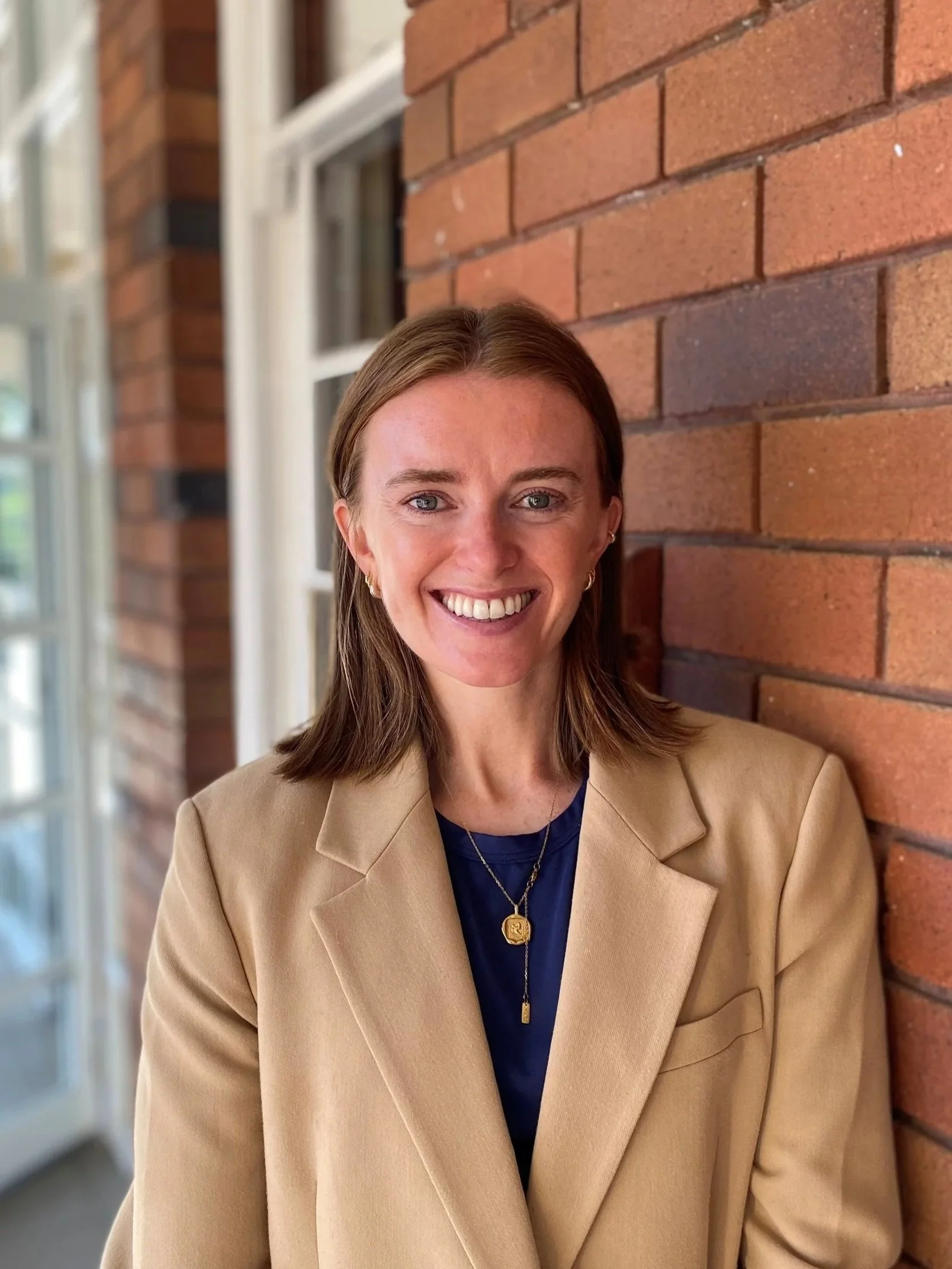 A woman with long brown hair and blue eyes smiling, standing against a brick wall with windows in the background, wearing a beige blazer and a navy top with a gold necklace.