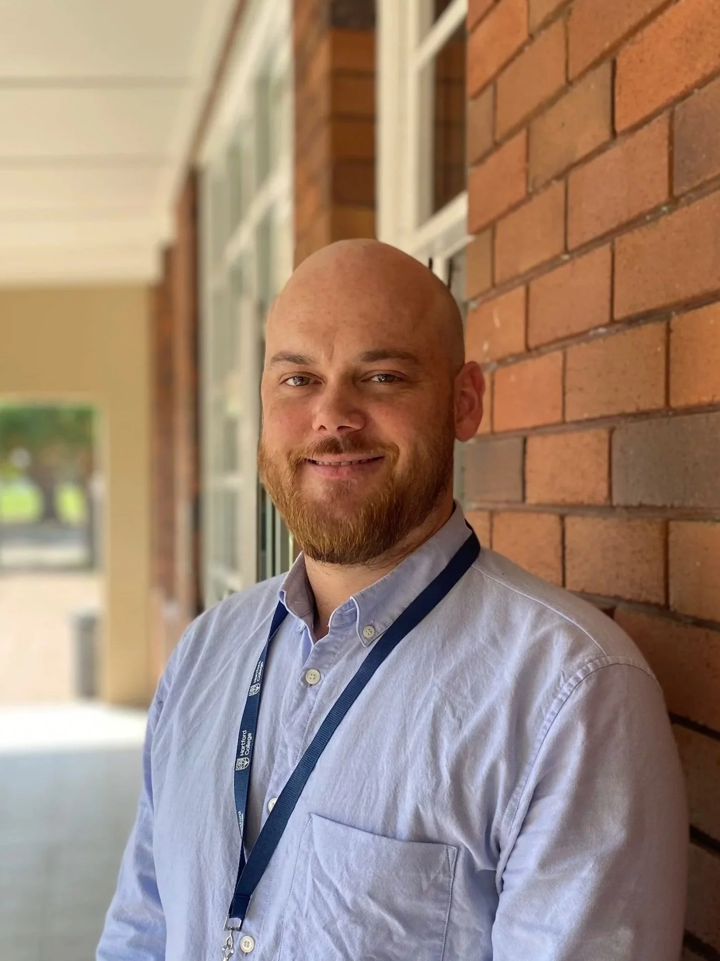 A man with a beard and a bald head smiling, standing outdoors next to a brick wall, wearing a light blue collared shirt and a dark lanyard.