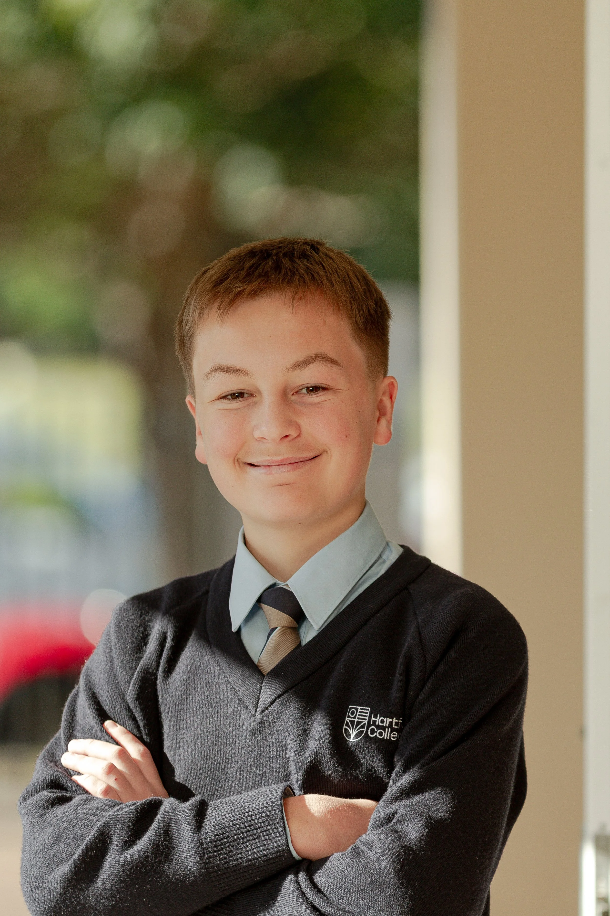 A teenage boy with short brown hair wearing a gray school uniform shirt, a striped tie, and a black sweater with a school logo, standing with arms crossed, smiling inside a school building.