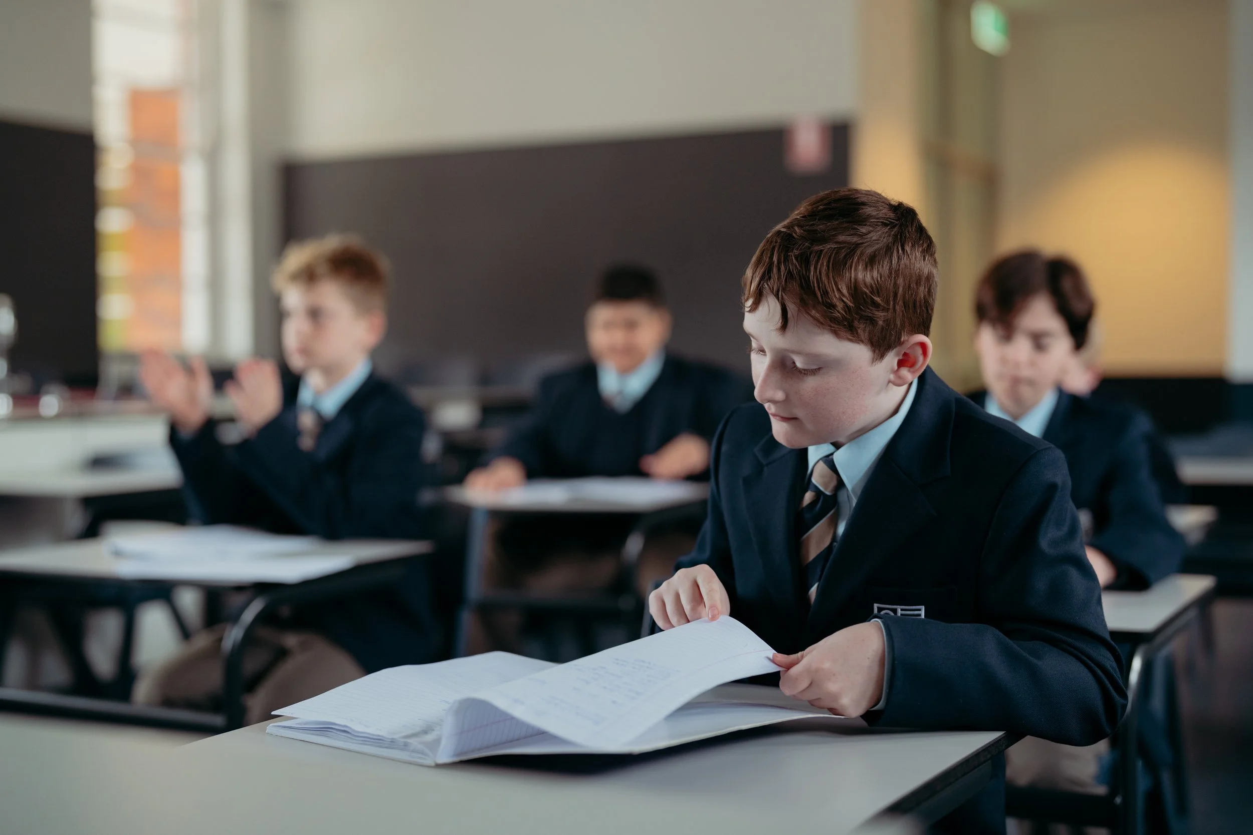 School classroom with young male students in formal uniforms, one student in foreground reading a paper, others clapping in the background.