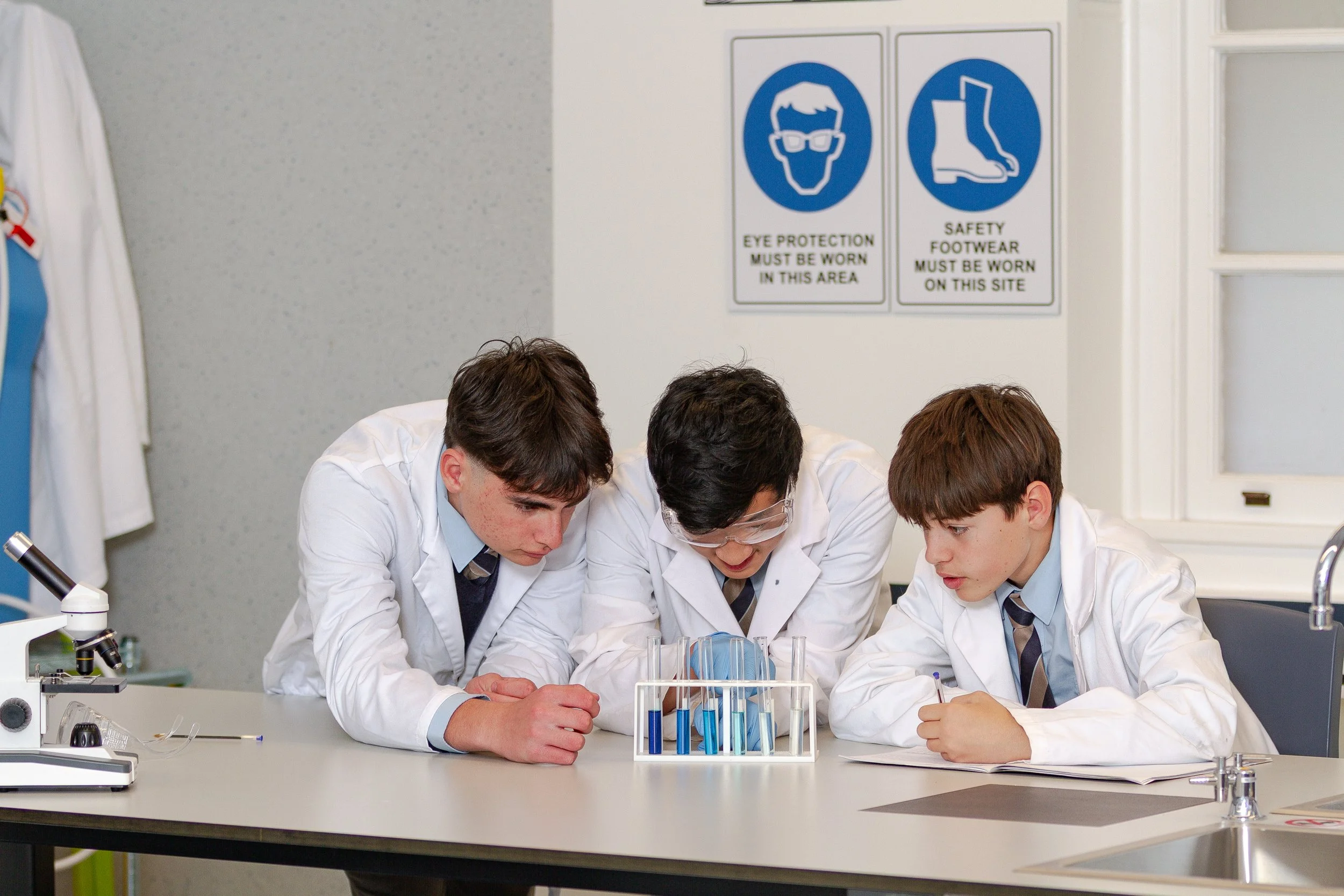 Three students in lab coats and ties working together at a science experiment on a classroom lab table, with science equipment including test tubes and microscopes present, and safety signs on the wall behind them.