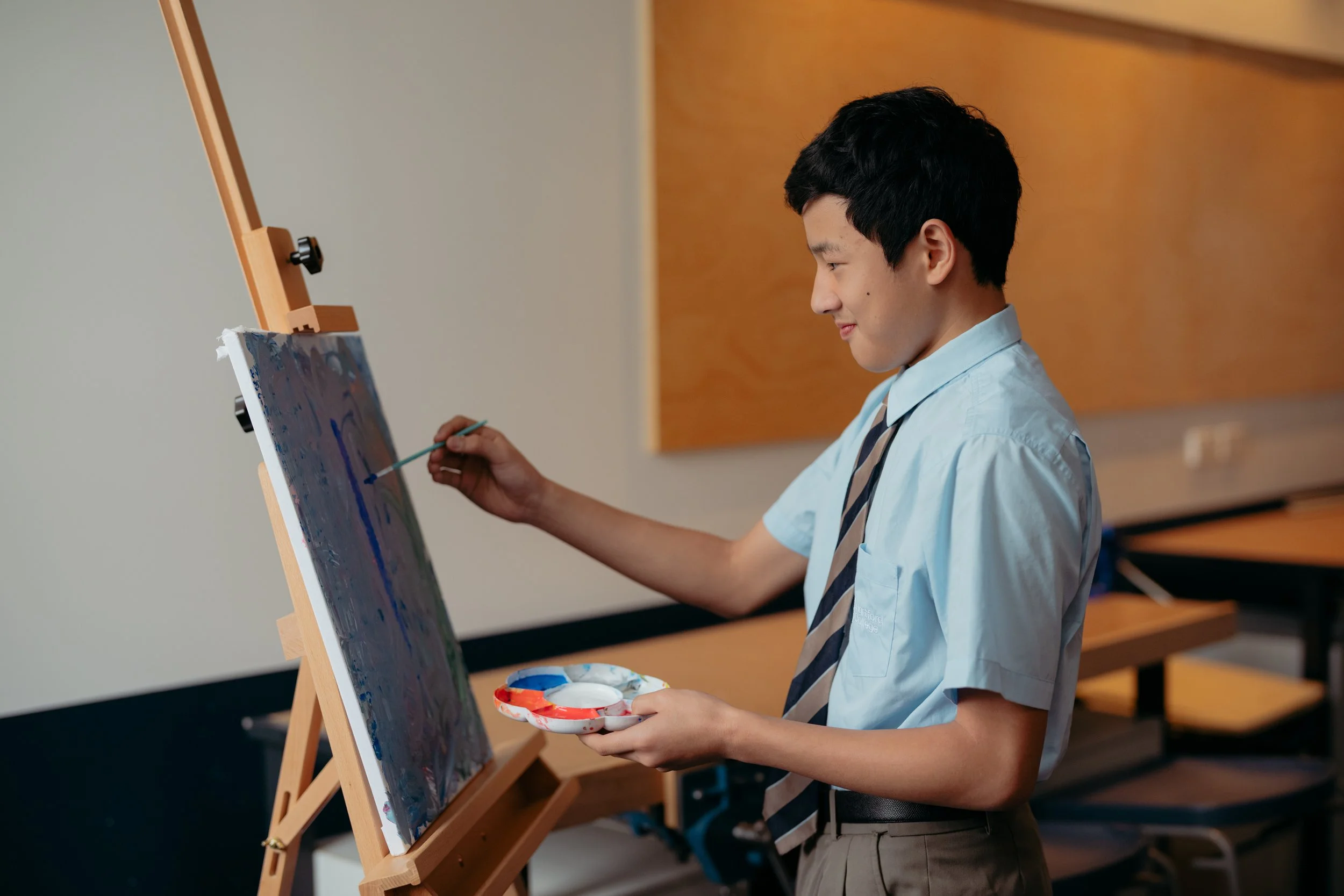 A young man in a school uniform painting on a canvas set up on an easel in a classroom or art studio.