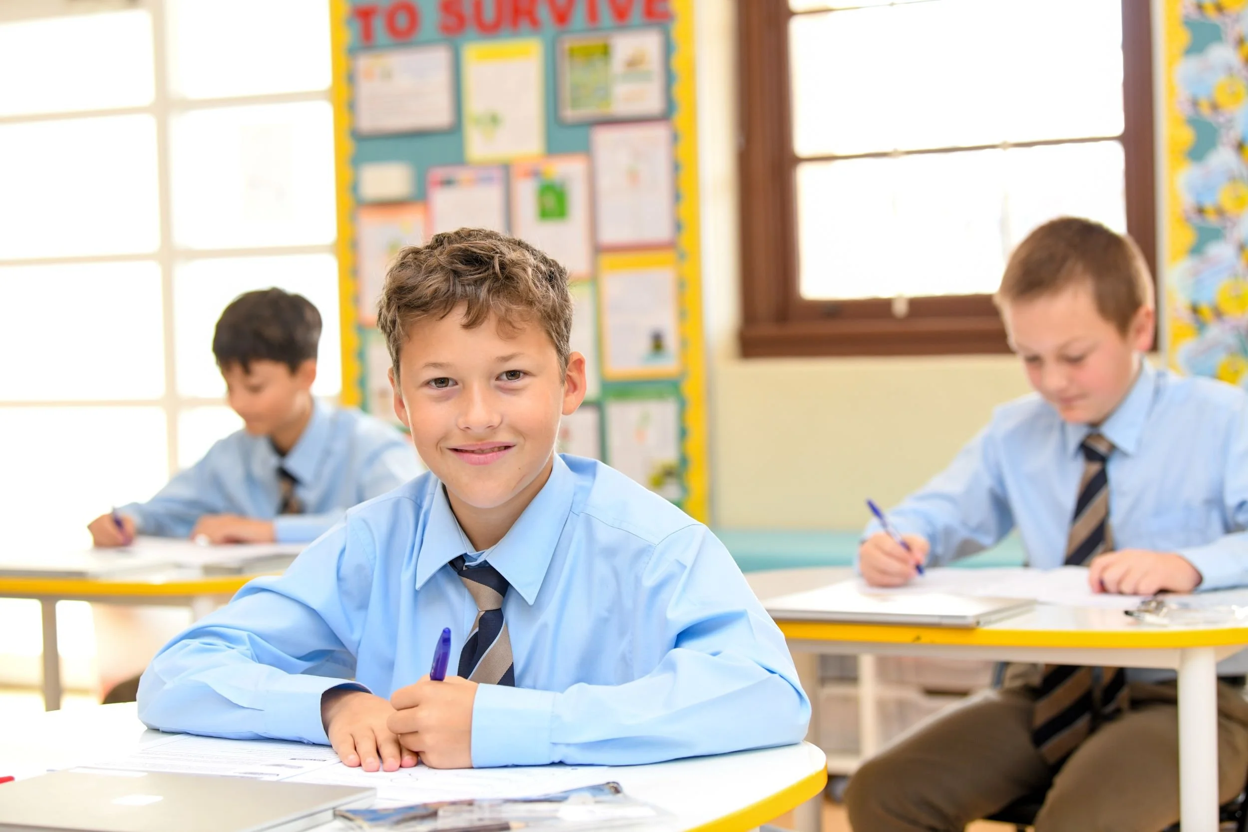 A classroom with three young boys in uniform sitting at desks. The focus is on a smiling boy with brown hair and blue eyes holding a purple pen. Other boys are visible in the background, engaged in writing. The classroom has large windows and colorful educational posters on the walls.
