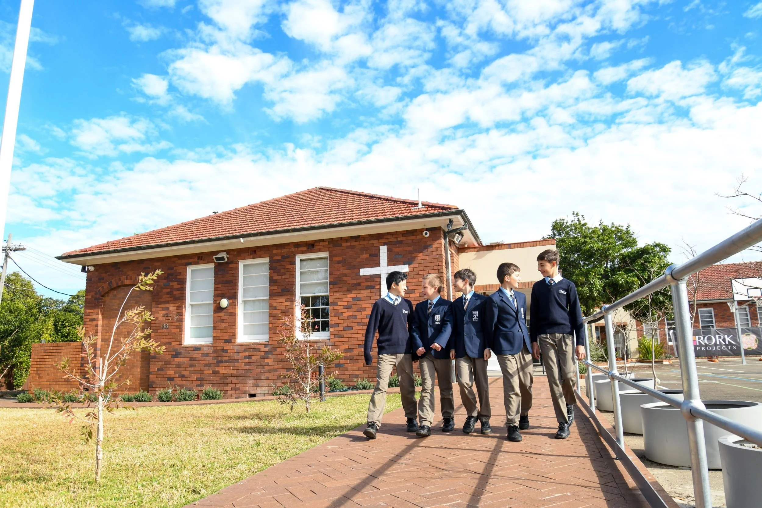 A group of school boys wearing uniforms walking outside next to a brick building with a cross on it, under a partly cloudy blue sky.