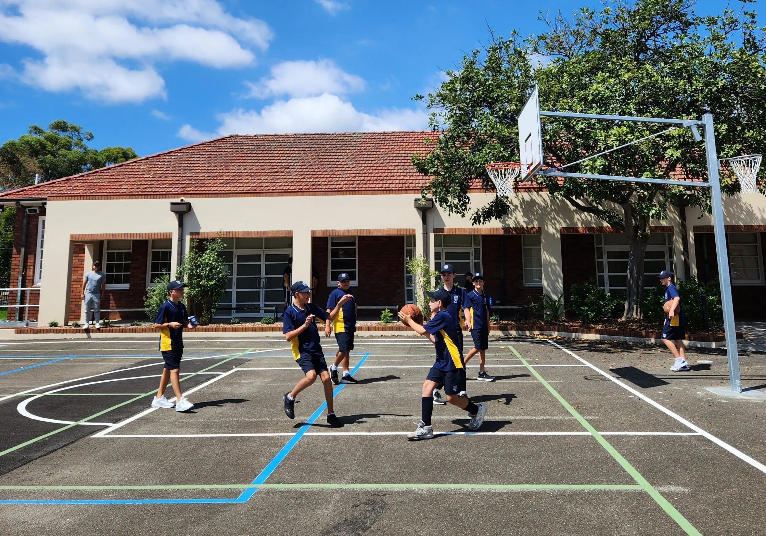 Children playing basketball on an outdoor court with a school building, basketball hoops, and trees under a blue sky with clouds.
