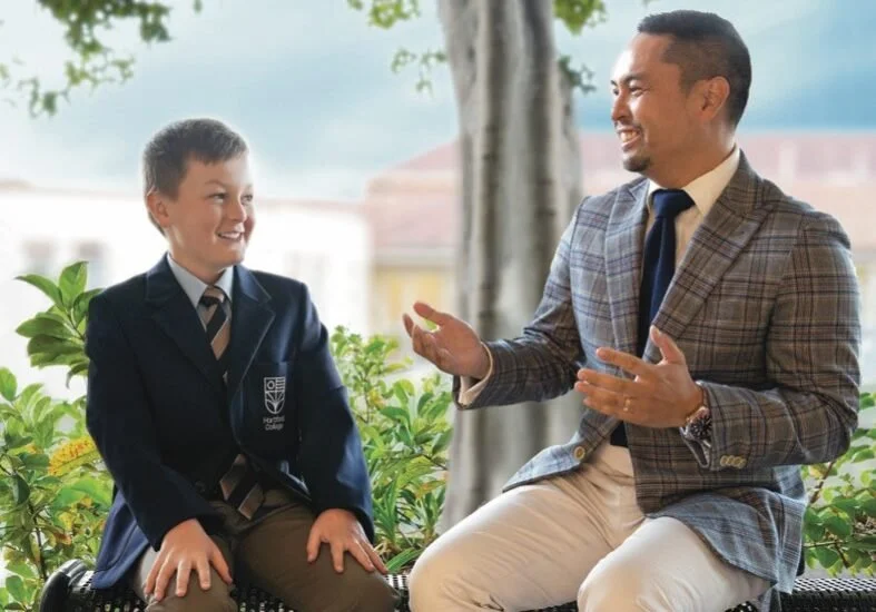 A boy and a teacher sitting outdoors, engaging in conversation and laughing, with greenery and trees in the background.