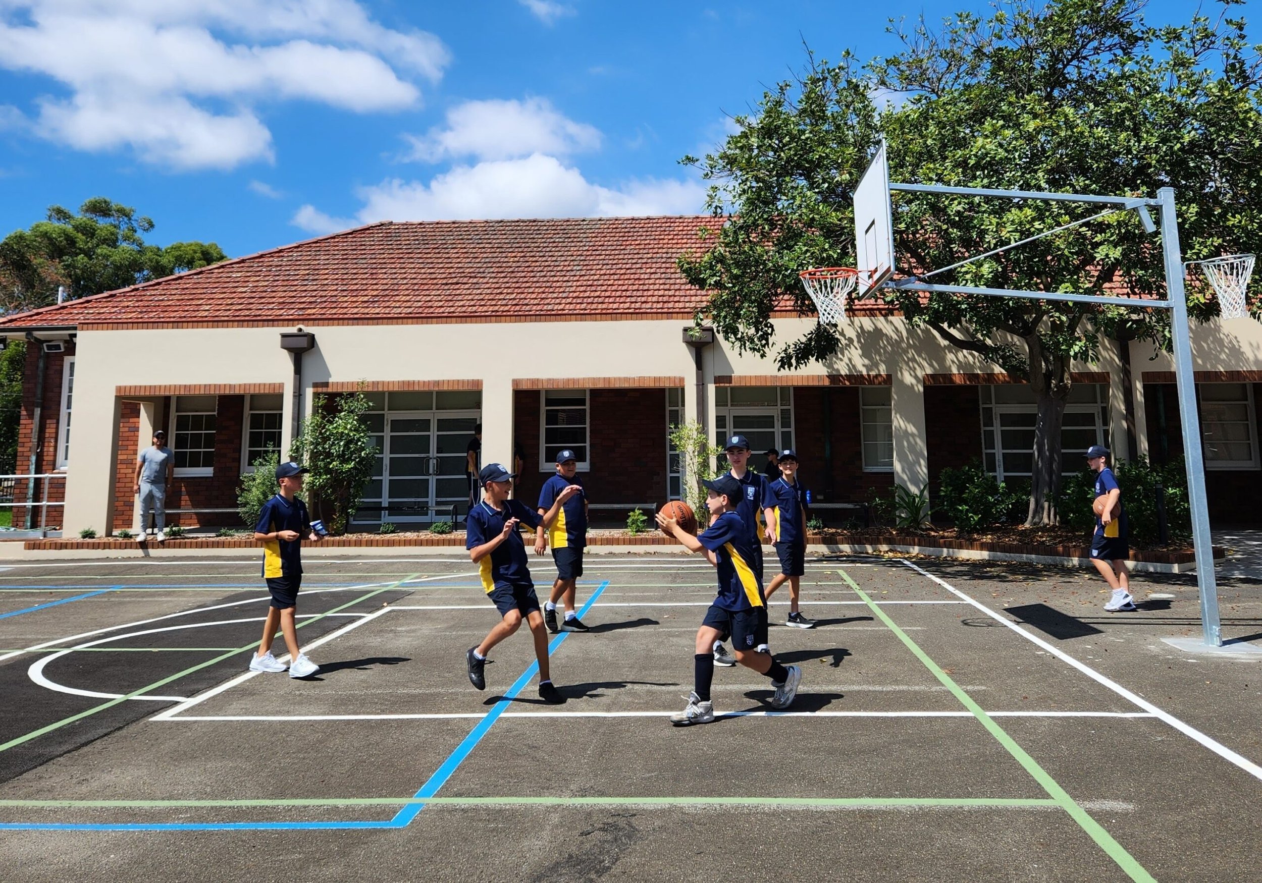 Children playing basketball on an outdoor court near a school building with a basketball hoop and a large tree, under a partly cloudy sky.
