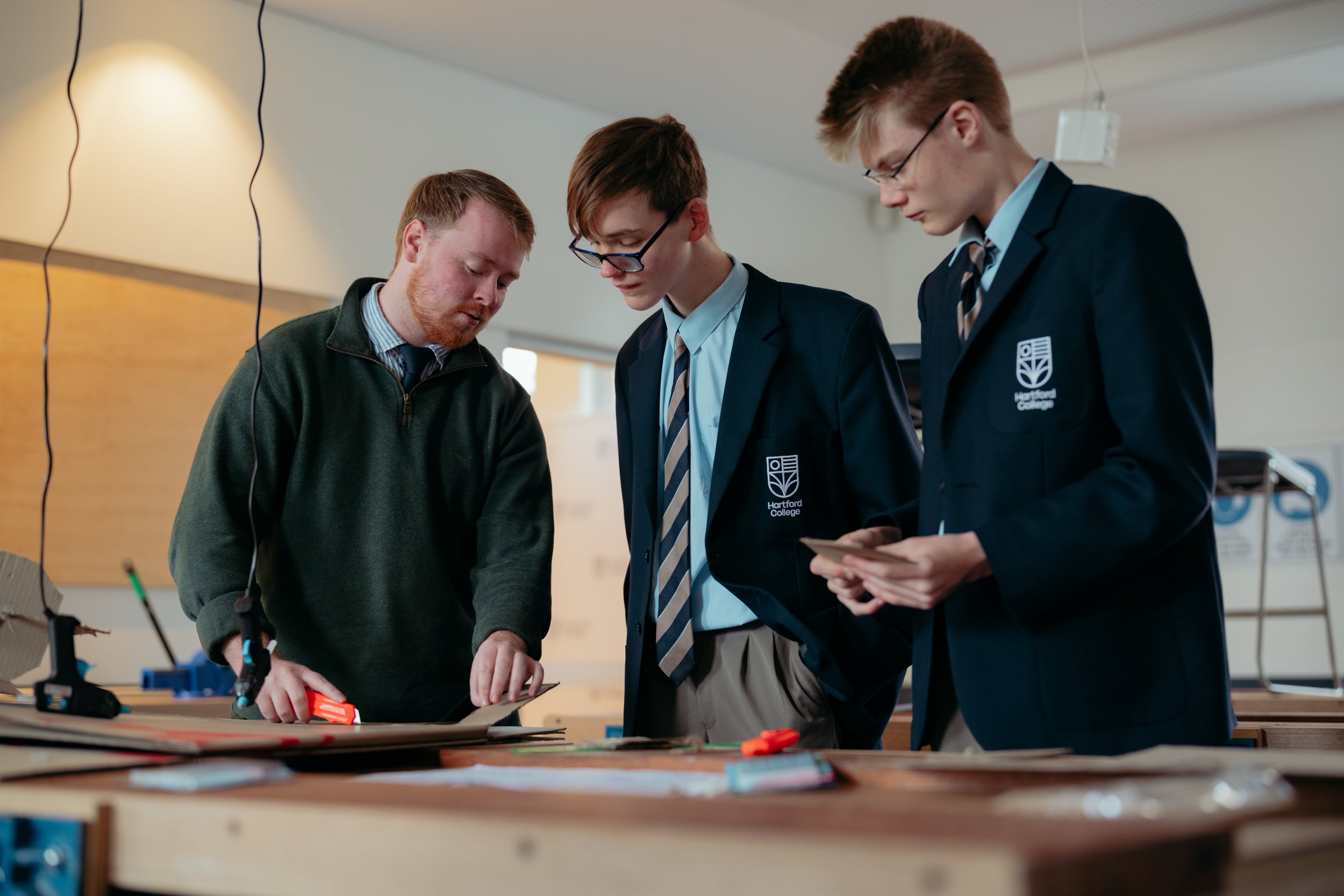 Three boys, two in school uniforms and one in casual clothes, are working together on a project at a table covered with tools and materials. They are looking down at their work in an indoor setting that appears to be a classroom or workshop.