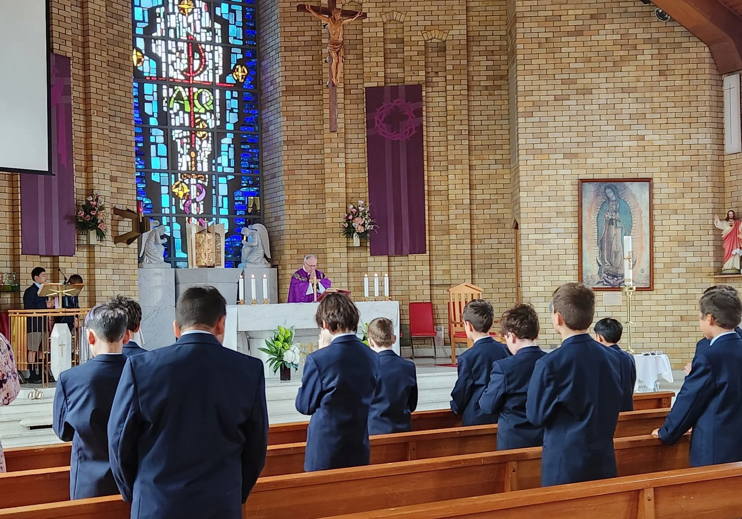 A church service with a young male choir in navy suits facing the altar. The priest, dressed in purple vestments, is standing at the altar, reading from a book. Behind him, a large stained glass window, a crucifix, and religious artwork are visible. There are pink floral arrangements and candles on the altar.