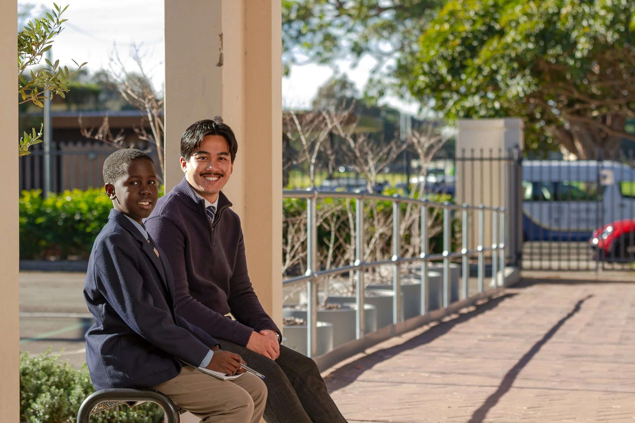 A young student in a school uniform and professional man with a mustache sitting on a bench at a schoolyard, smiling with a background of trees, fence, and parked cars.