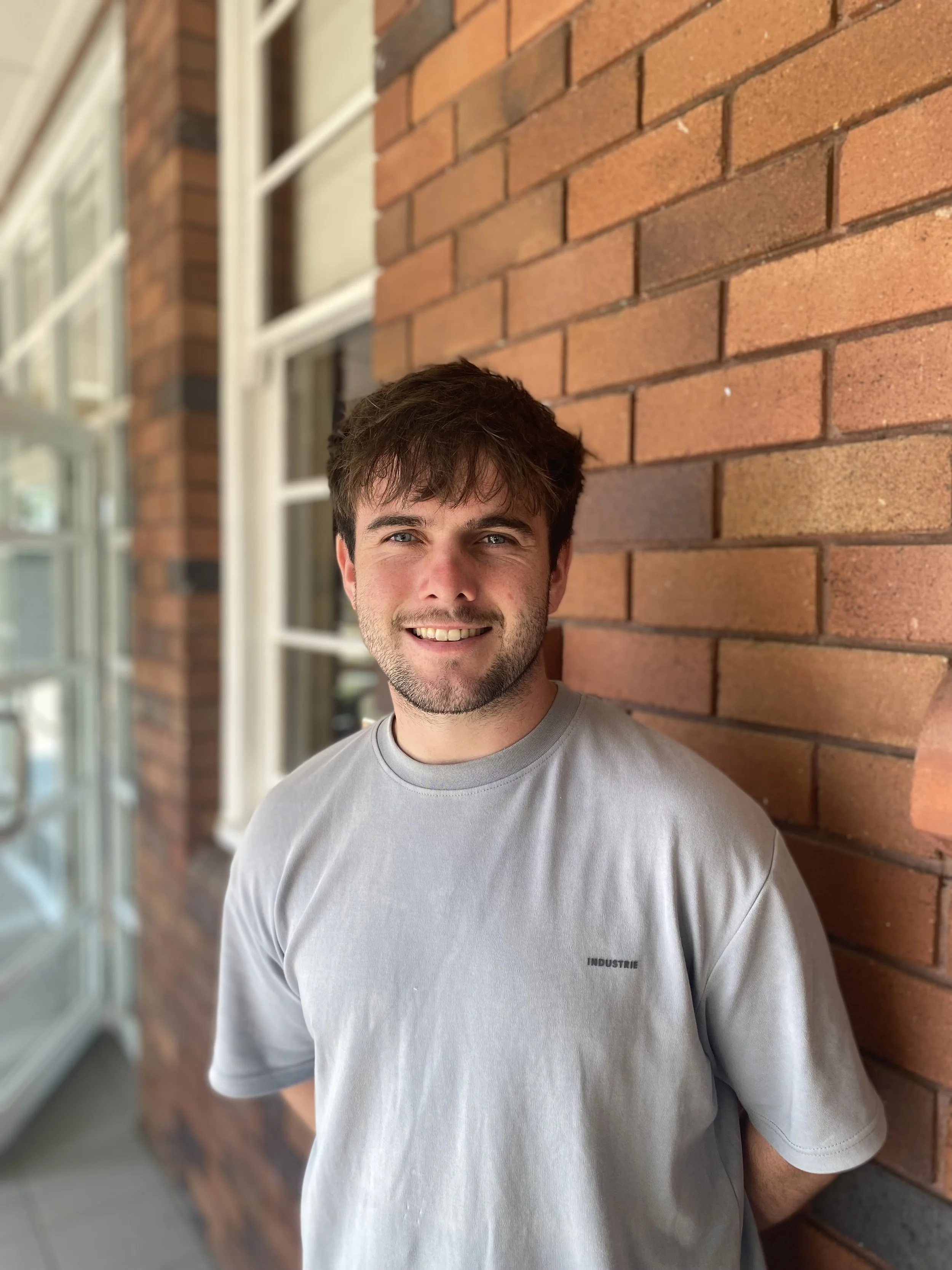 A man with brown hair and a light beard smiling while leaning against a red brick wall outdoors, wearing a light gray T-shirt with the word 'INDUSTRIE' printed on it.