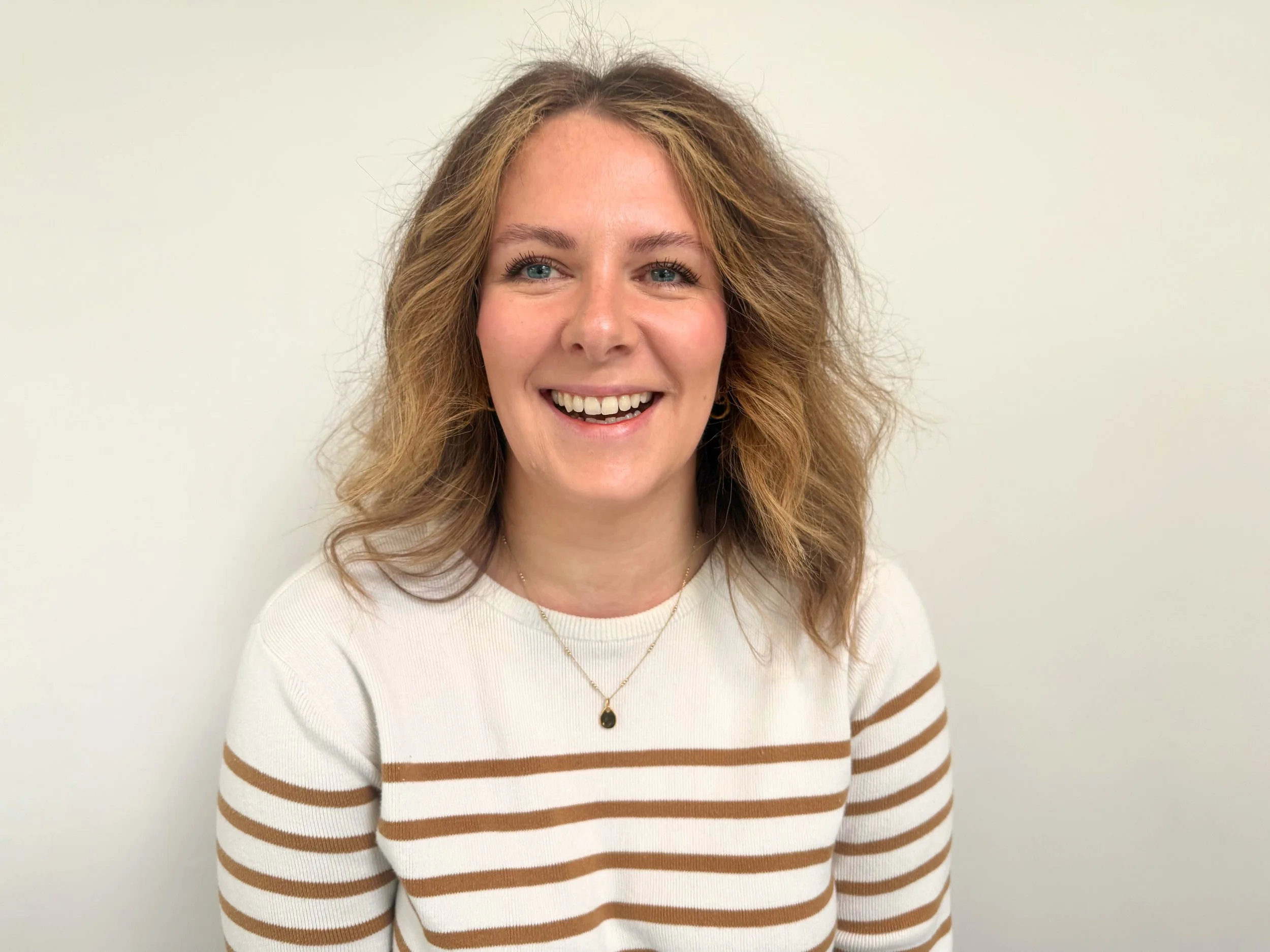 A woman with wavy, shoulder-length hair, and blue eyes, smiling, wearing a white and brown striped sweater, and a necklace with a black pendant, standing against a plain off-white background.