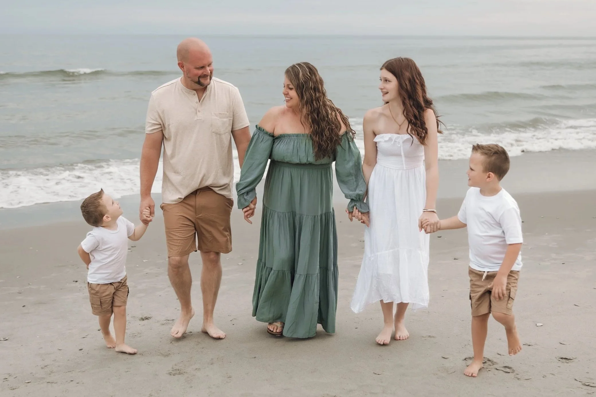 A family walking on the beach holding hands, with the ocean and waves in the background. The family includes two children and three adults, all smiling and enjoying their time at the beach.