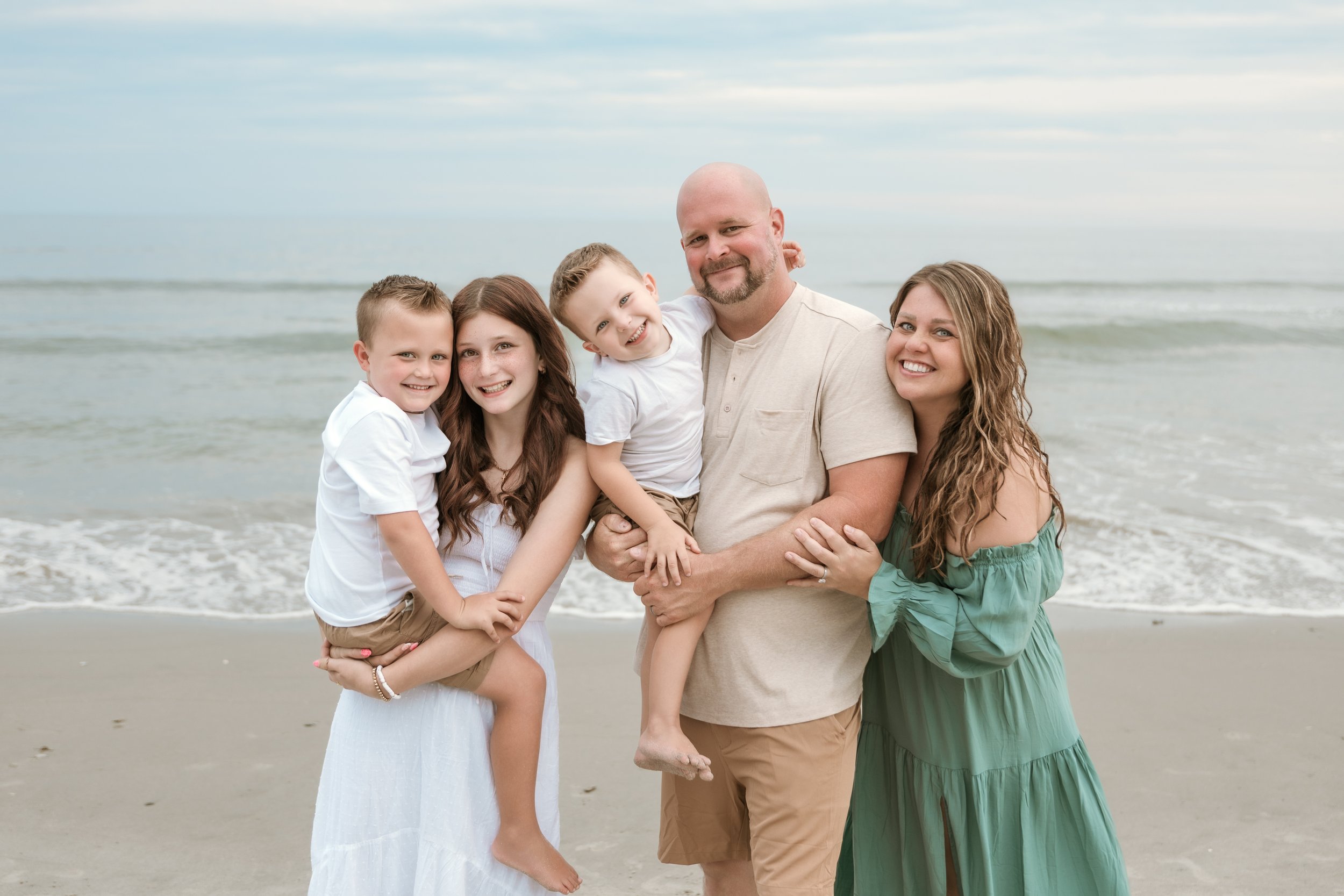 A family of five standing on a beach, smiling at the camera. The father is holding a young boy, and two girls are standing next to him, with the mother on the other side, all standing close together.
