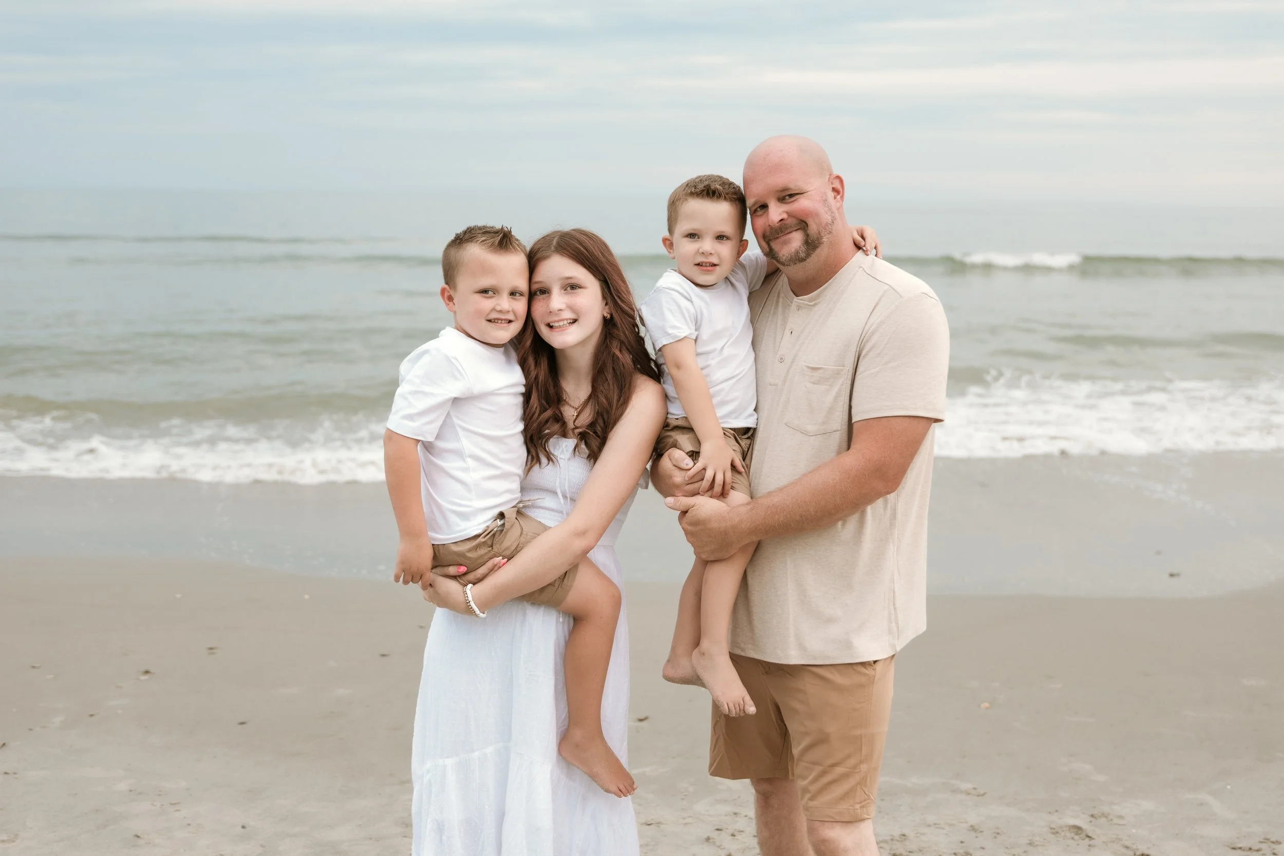 A family of five, including a mother, father, and three young children, standing on a beach near the ocean, smiling at the camera.