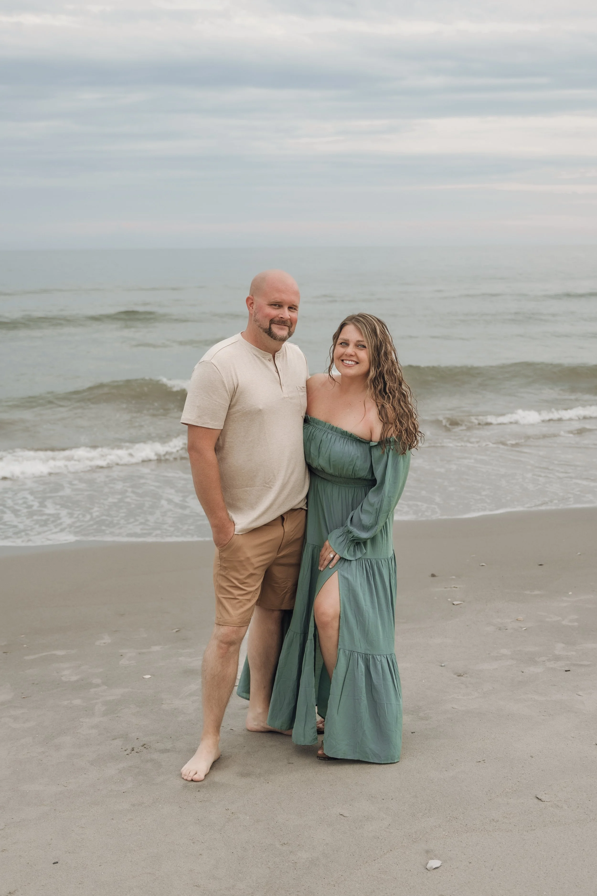 A couple standing on the beach with ocean waves in the background, the man with a beard and bald head wearing a beige shirt and tan shorts, and the woman with long curly hair wearing a green off-shoulder dress, both smiling.