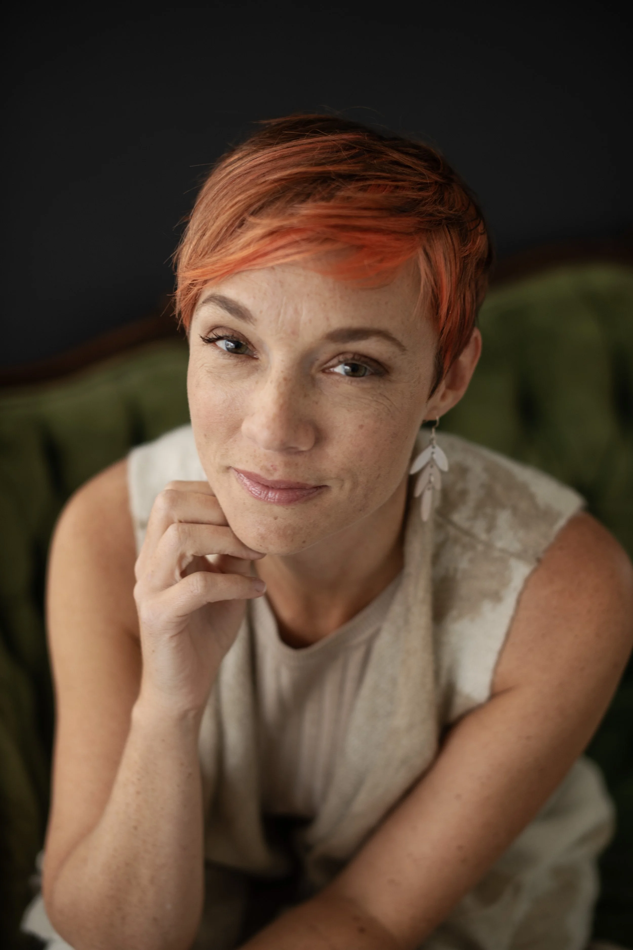 Close-up portrait of a woman with short, red hair, freckles, and wearing white leaf-shaped earrings, sitting on a green velvet chair with a dark background.